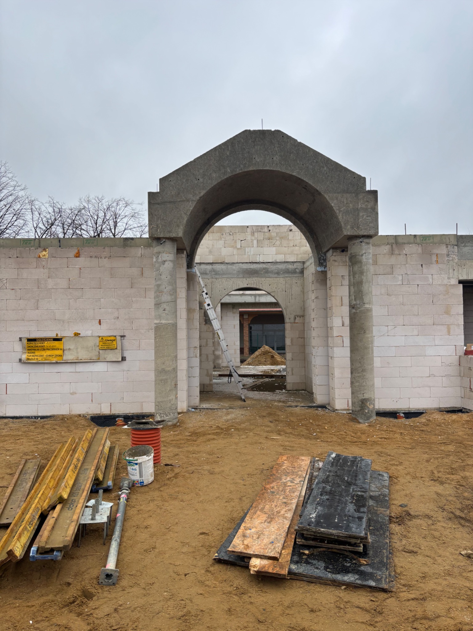 Surowy stan domu w trakcie budowy z białych bloczków, widoczne betonowe łuki wejściowe, drabina oparta o ścianę, oraz materiały budowlane na piaszczystym podłożu.