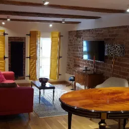 Salon z ceglaną ścianą, żółtymi okiennicami i drewnianymi belkami stropowymi, oświetlony punktowo, z czerwonym narożnikiem i ciemnymi, lakierowanymi meblami.