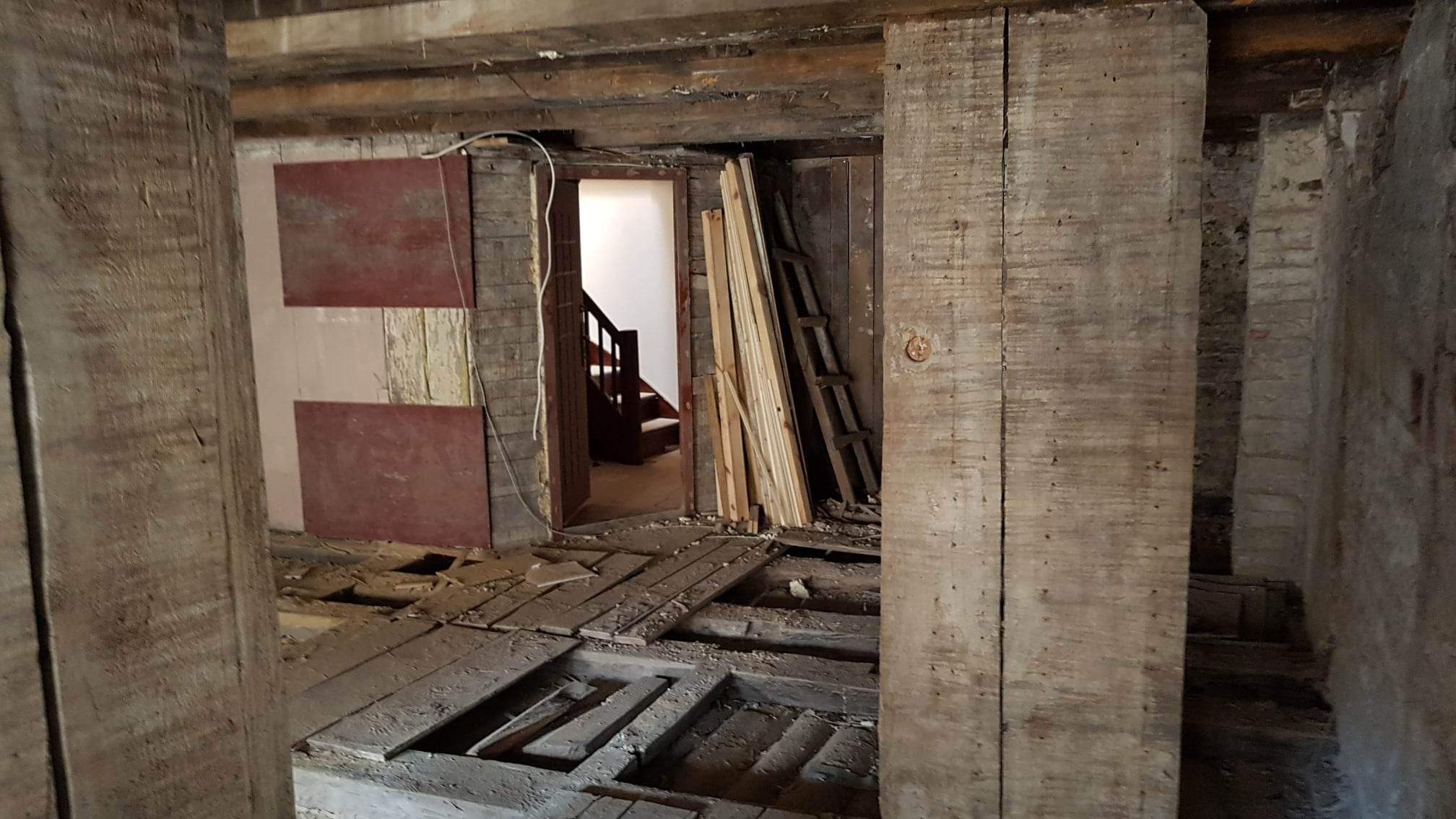 Wnętrze starego domu w trakcie rozbiórki lub gruntownego remontu, widoczne odsłonięte belki stropowe, fragmenty podłogi, ściany z cegły i desek oraz schody w głębi