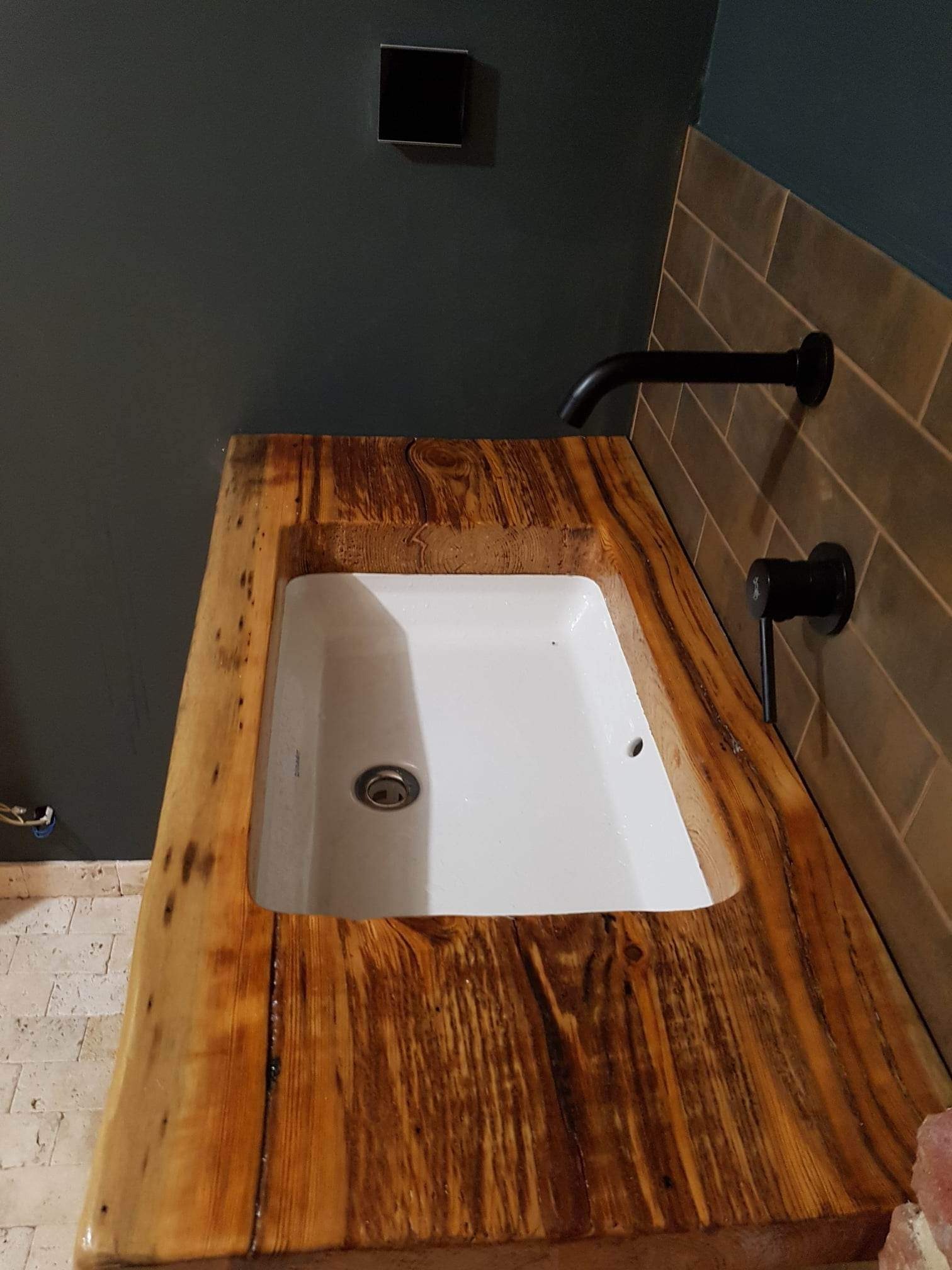Biała umywalka wbudowana w blat z surowego, lakierowanego drewna z widocznymi słojami i sękami, czarna bateria ścienna, szare płytki imitujące cegłę na ścianie, ciemnoszara ściana w tle.
