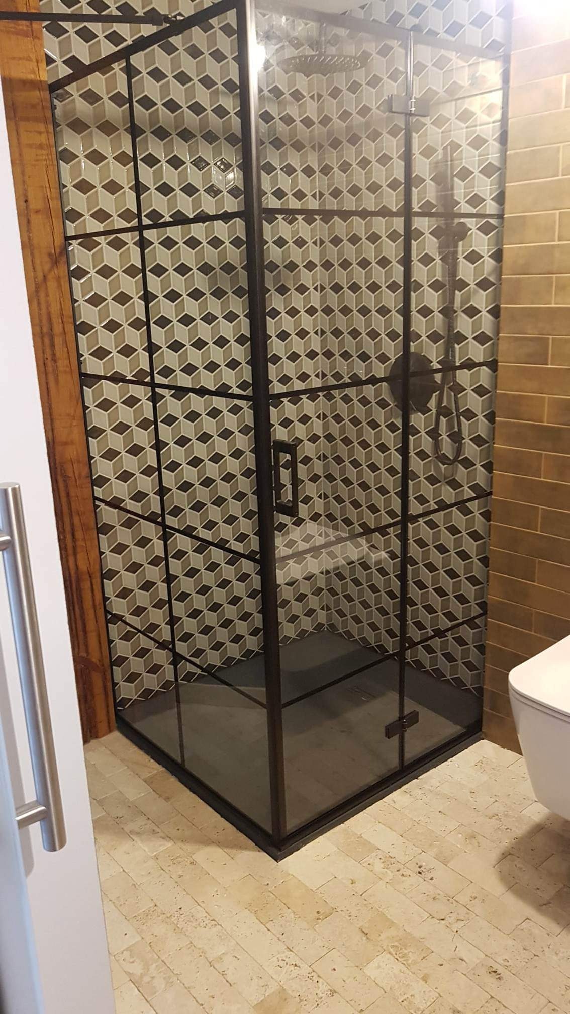 Kabina prysznicowa z czarną ramą i geometrycznymi płytkami w odcieniach beżu i brązu, widoczna część toalety, podłoga wyłożona jasnymi, podłużnymi płytkami imitującymi kamień.
