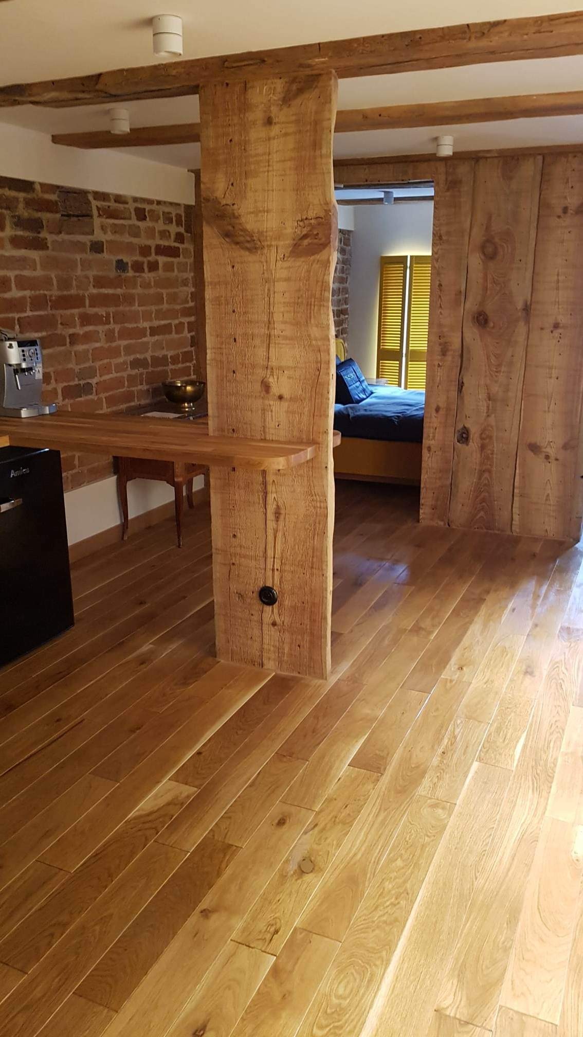 Rustykalne wykończenie wnętrza z drewnianą kolumną, blatem barowym, parkietem i ścianą z czerwonej cegły, widok na sypialnię z żółtymi żaluzjami.