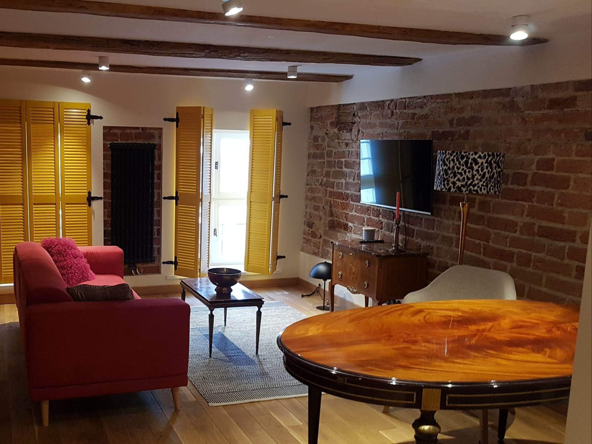 Salon z ceglaną ścianą, żółtymi okiennicami i drewnianymi belkami stropowymi, oświetlony punktowo, z czerwonym narożnikiem i ciemnymi, lakierowanymi meblami.