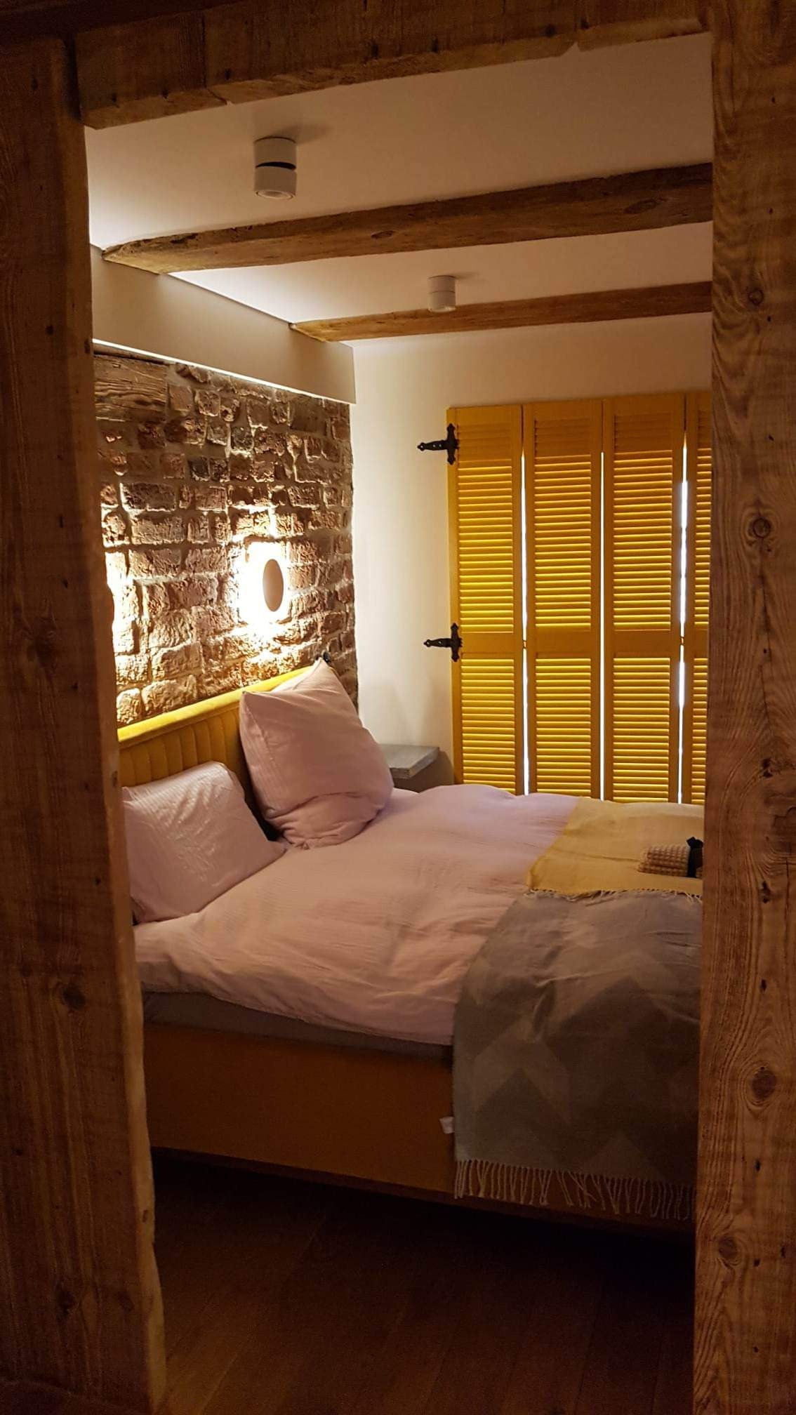 Rustykalna sypialnia z ceglaną ścianą, żółtymi okiennicami, drewnianymi belkami stropowymi i łóżkiem z różową pościelą, widziana zza drewnianej ramy drzwi.