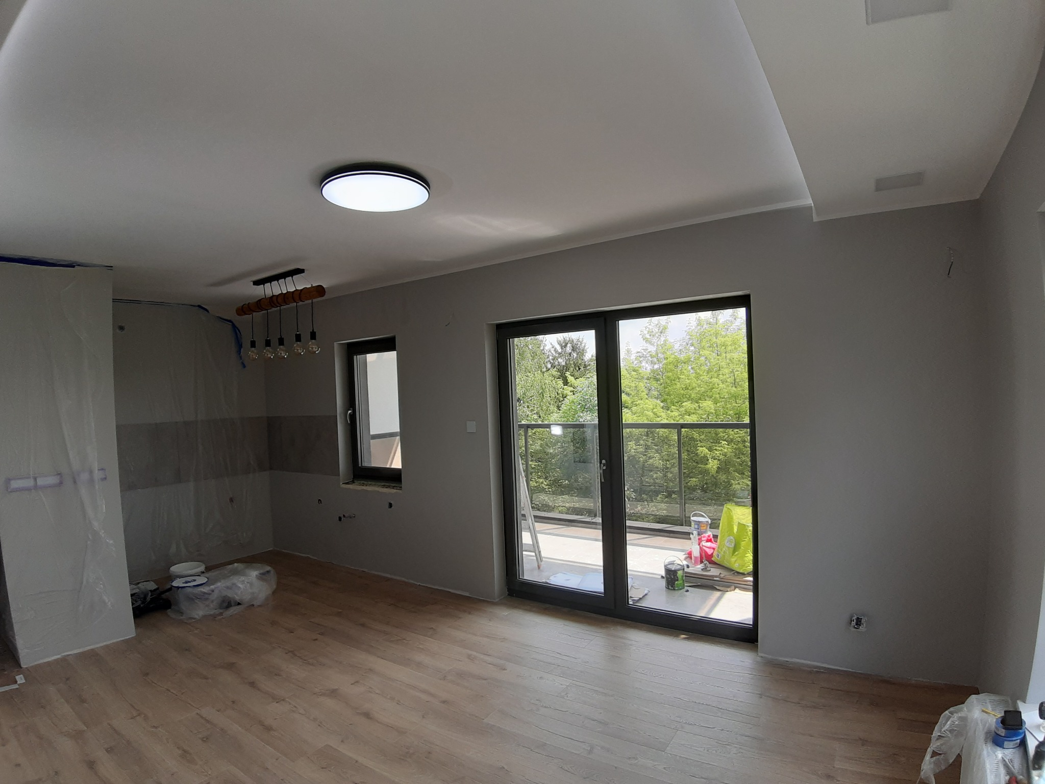 Pokój w trakcie prac wykończeniowych: świeżo położona drewniana podłoga, szare ściany, zakryte folią meble kuchenne, widok na balkon z zielenią w tle.