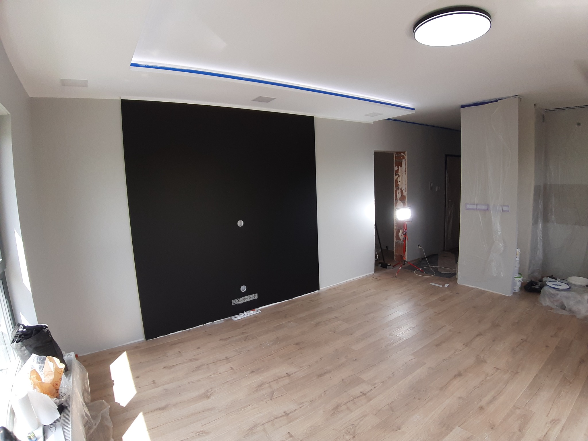 Salon w trakcie prac wykończeniowych: ułożona podłoga z paneli, ściany częściowo pomalowane na szaro i czarno, sufit z oświetleniem LED zabezpieczony taśmą malarską, widoczne narzędzia i materiały...