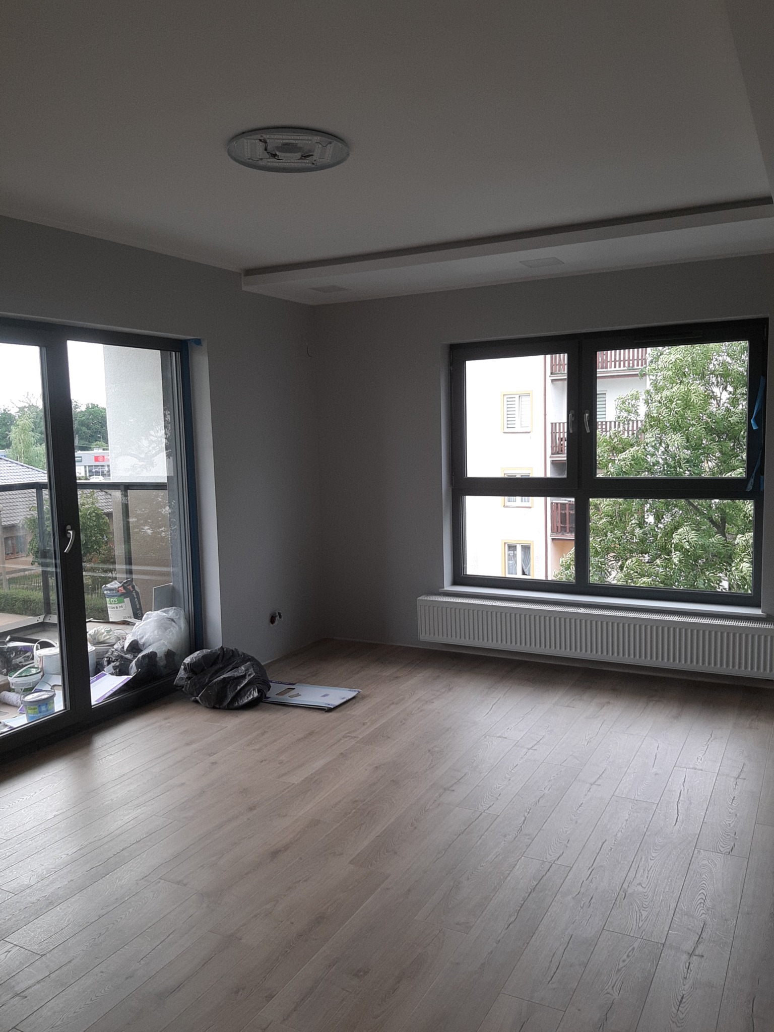 Pusta przestrzeń w nowym mieszkaniu z panelami podłogowymi, grzejnikiem pod oknem i workami z materiałami budowlanymi przy oknie balkonowym.