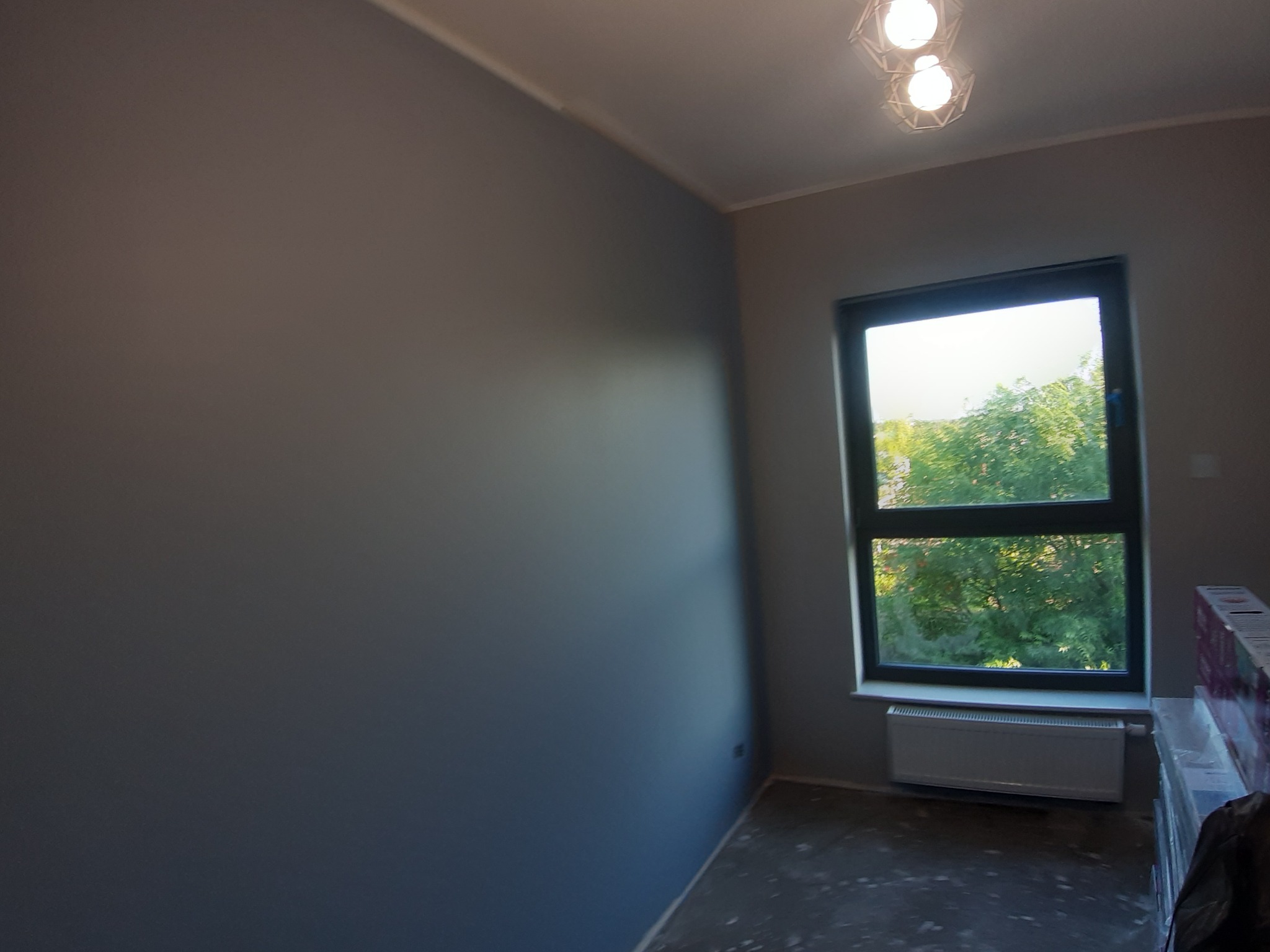 Świeżo pomalowane na szaro wnętrze pokoju z dużym oknem i grzejnikiem, widoczne przygotowane materiały budowlane w rogu, nowoczesna lampa sufitowa.