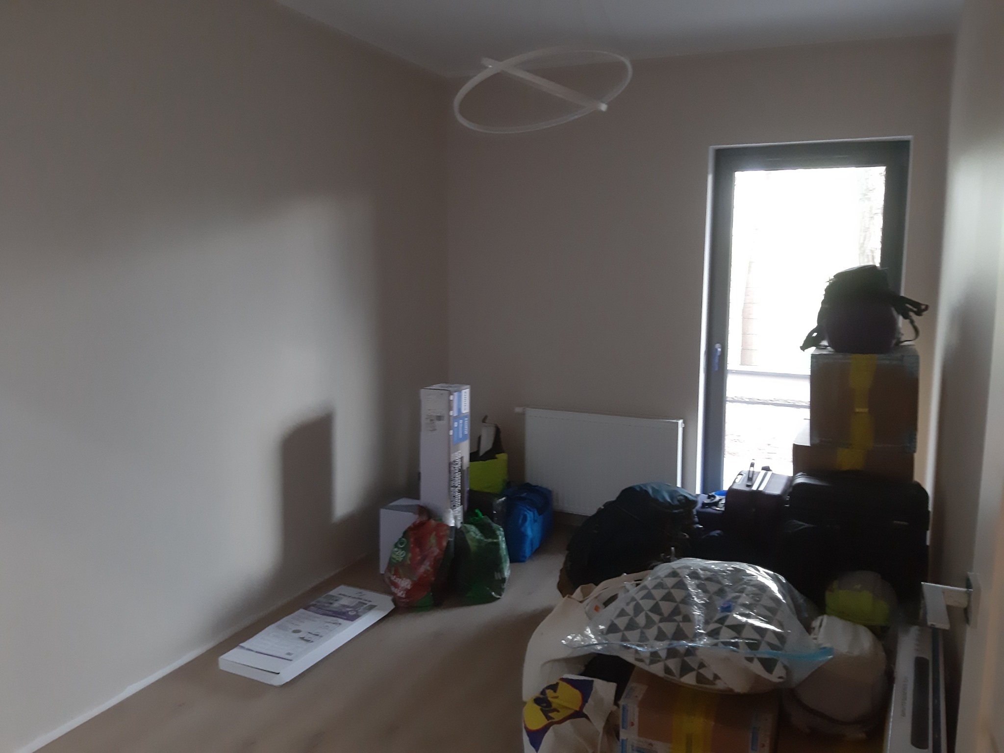 Pusty pokój z jasnymi ścianami i drewnianą podłogą, zagracony torbami podróżnymi, kartonami i innymi przedmiotami, lampa sufitowa w kształcie dwóch przecinających się okręgów, okno z widokiem...