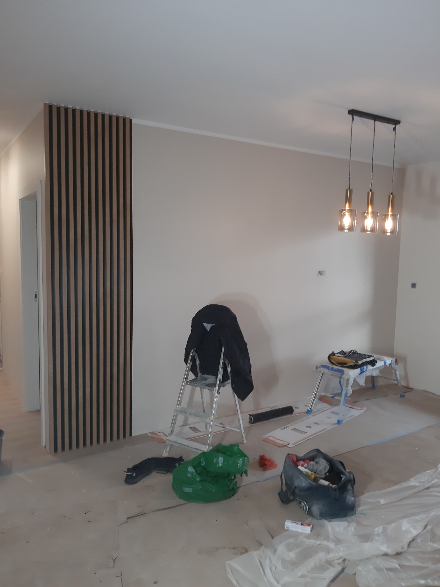 Pokój w trakcie prac wykończeniowych: pionowe lamele dekoracyjne na ścianie, drabina z kurtką, lampa wisząca z trzema szklanymi kloszami, podłoga zabezpieczona folią malarską.