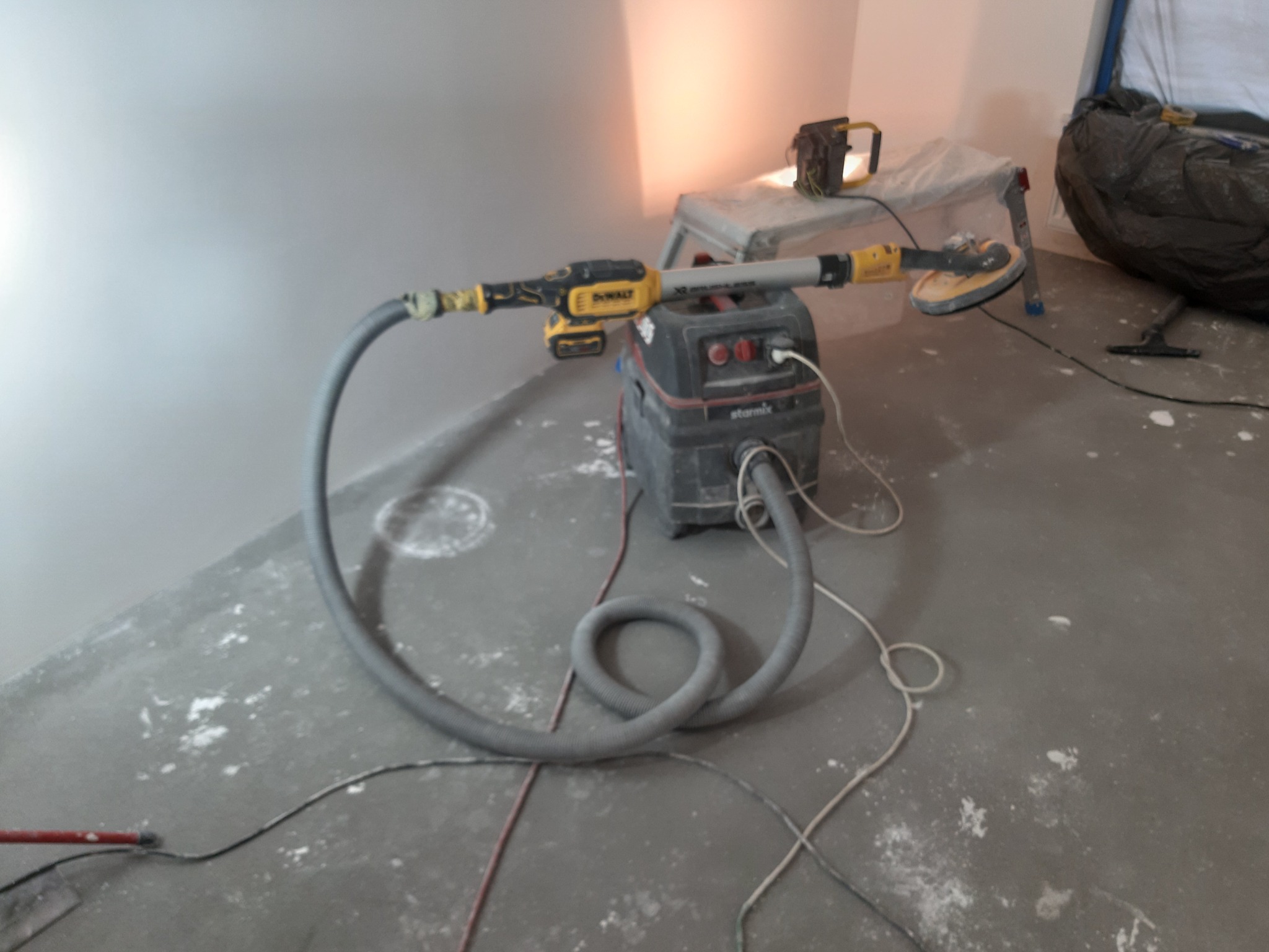 Szlifierka do gładzi gipsowych DeWalt podłączona do odkurzacza Starmix podczas pracy w pomieszczeniu, widoczne ślady gipsu na podłodze.