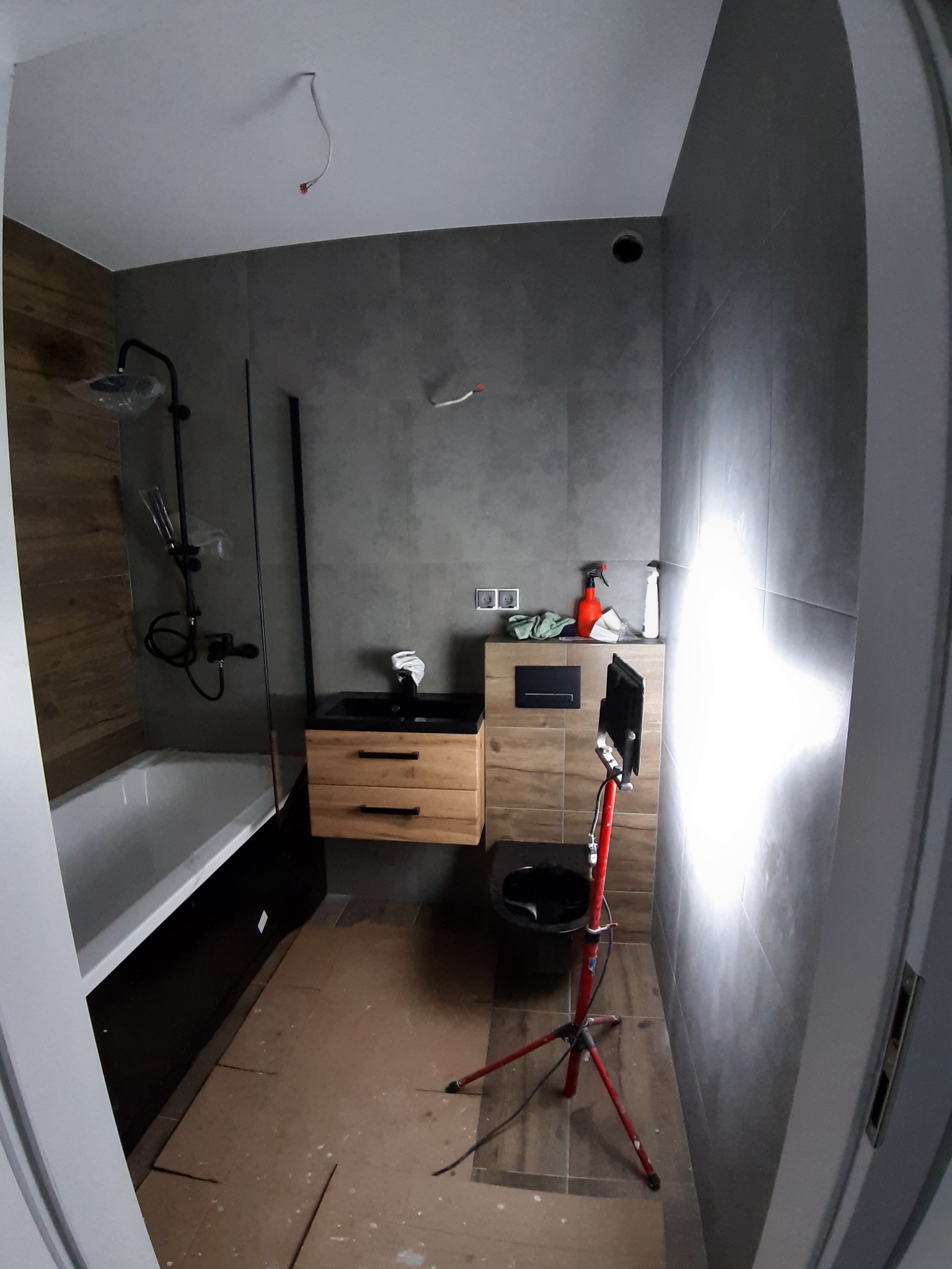 Wnętrze łazienki w trakcie wykańczania, widoczna wanna z czarną obudową, umywalka z drewnianą szafką, toaleta, szare płytki na ścianach, lampa budowlana na statywie, częściowo zabezpieczona podłoga.