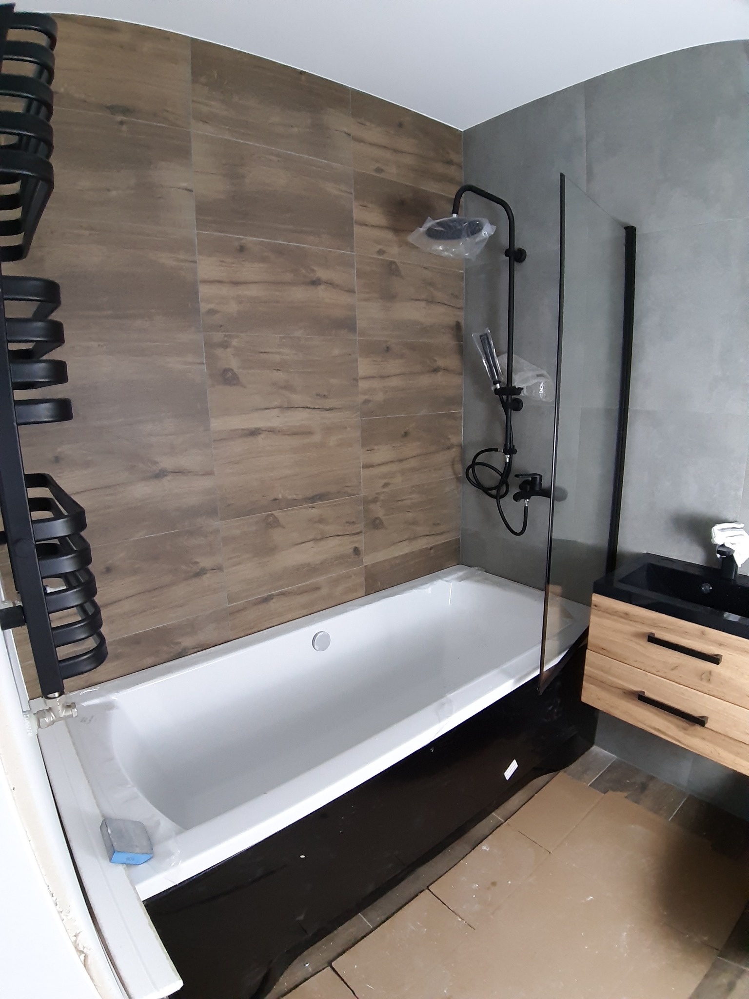 Nowoczesna łazienka w trakcie wykańczania: biała wanna z czarnym obramowaniem, czarna armatura prysznicowa w folii ochronnej, drewnopodobne płytki na ścianie i szara ściana, szafka pod umywalką...