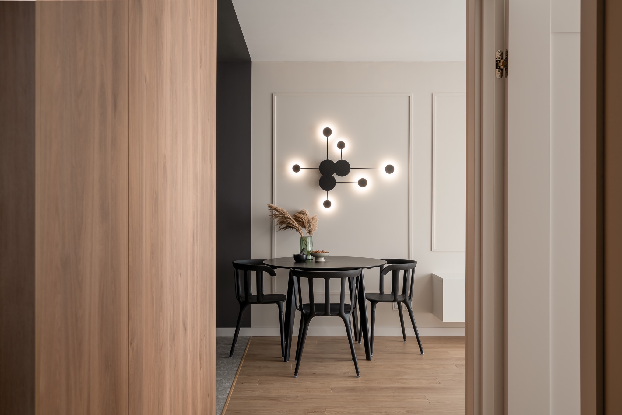 Minimalistyczna jadalnia z okrągłym czarnym stołem i krzesłami, ozdobiona modernistyczną lampą ścienną i suszonymi trawami w szklanym wazonie.
