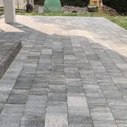 Ułożona szara kostka betonowa tworząca taras przy domu, z niskim stopniem z ciemniejszej kostki. W tle widoczne drzewa i trawa.