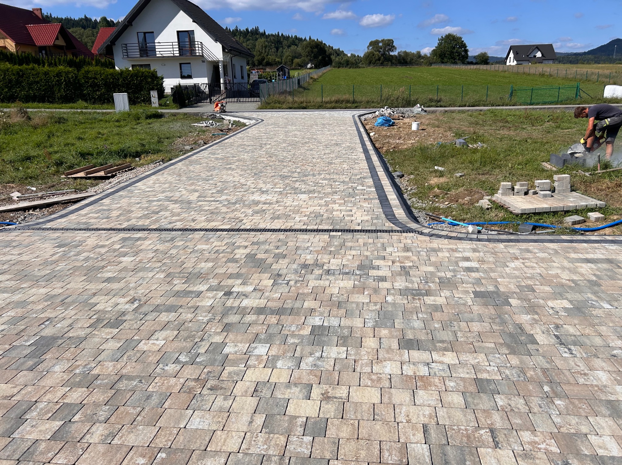 Szeroki podjazd z wielobarwnej kostki brukowej prowadzący do domu, z widocznym pracownikiem docinającym kostkę w tle.