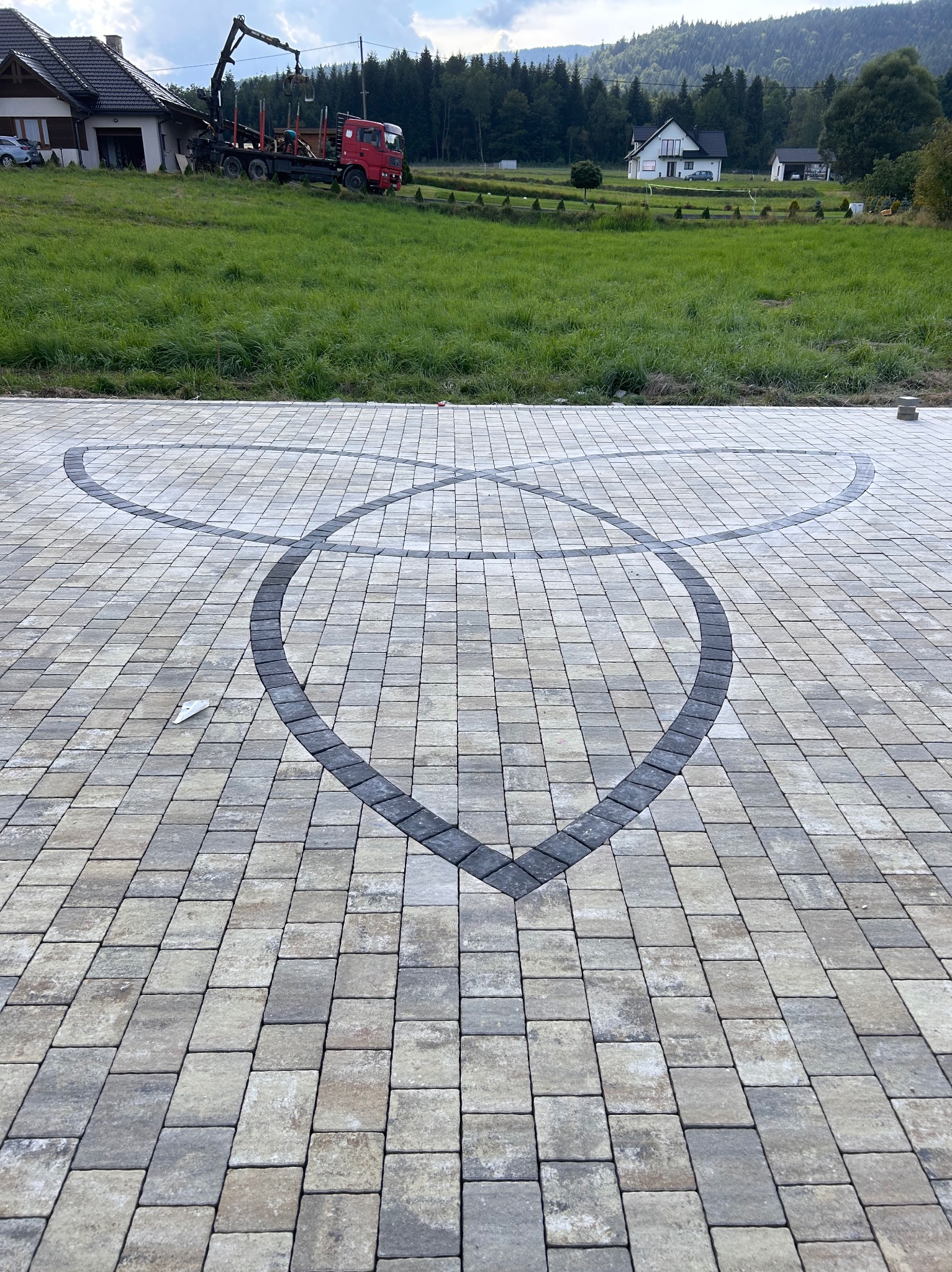 Ułożona kostka brukowa w odcieniach szarości i beżu z dekoracyjnym wzorem z ciemniejszych kostek tworzących dwa przecinające się półkola na tle zielonego trawnika i zabudowań.