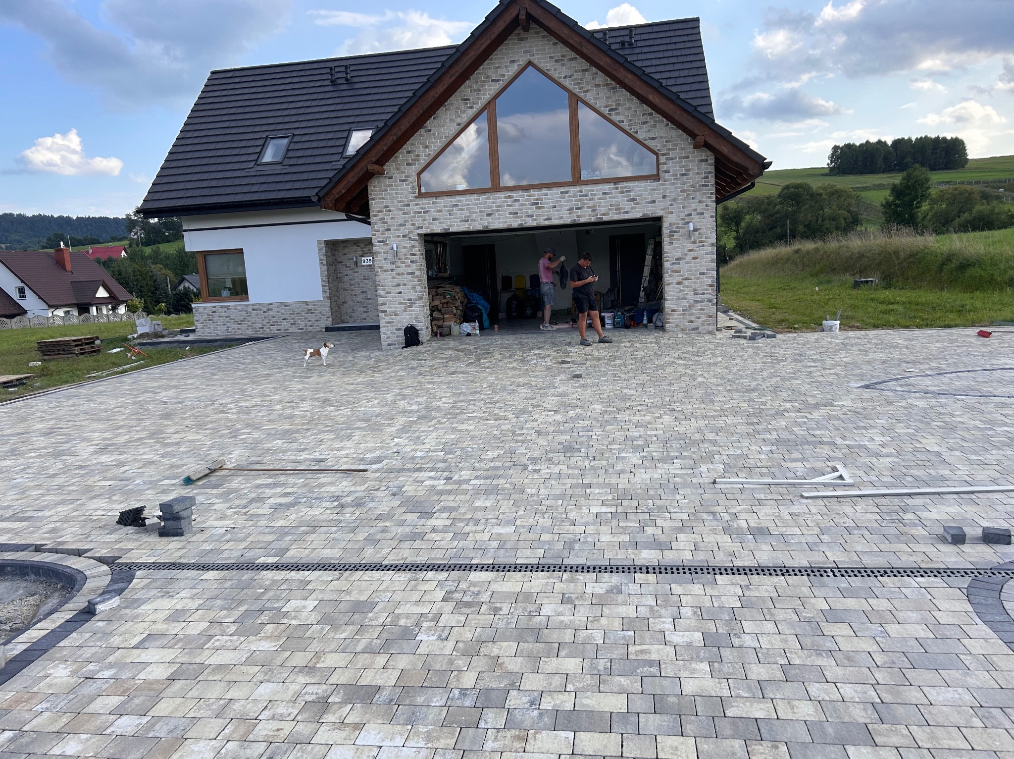 Ułożona kostka brukowa w odcieniach szarości na podjeździe przed nowoczesnym domem z garażem, widoczne elementy wykończeniowe i pracownicy w garażu, mały piesek na podjeździe.