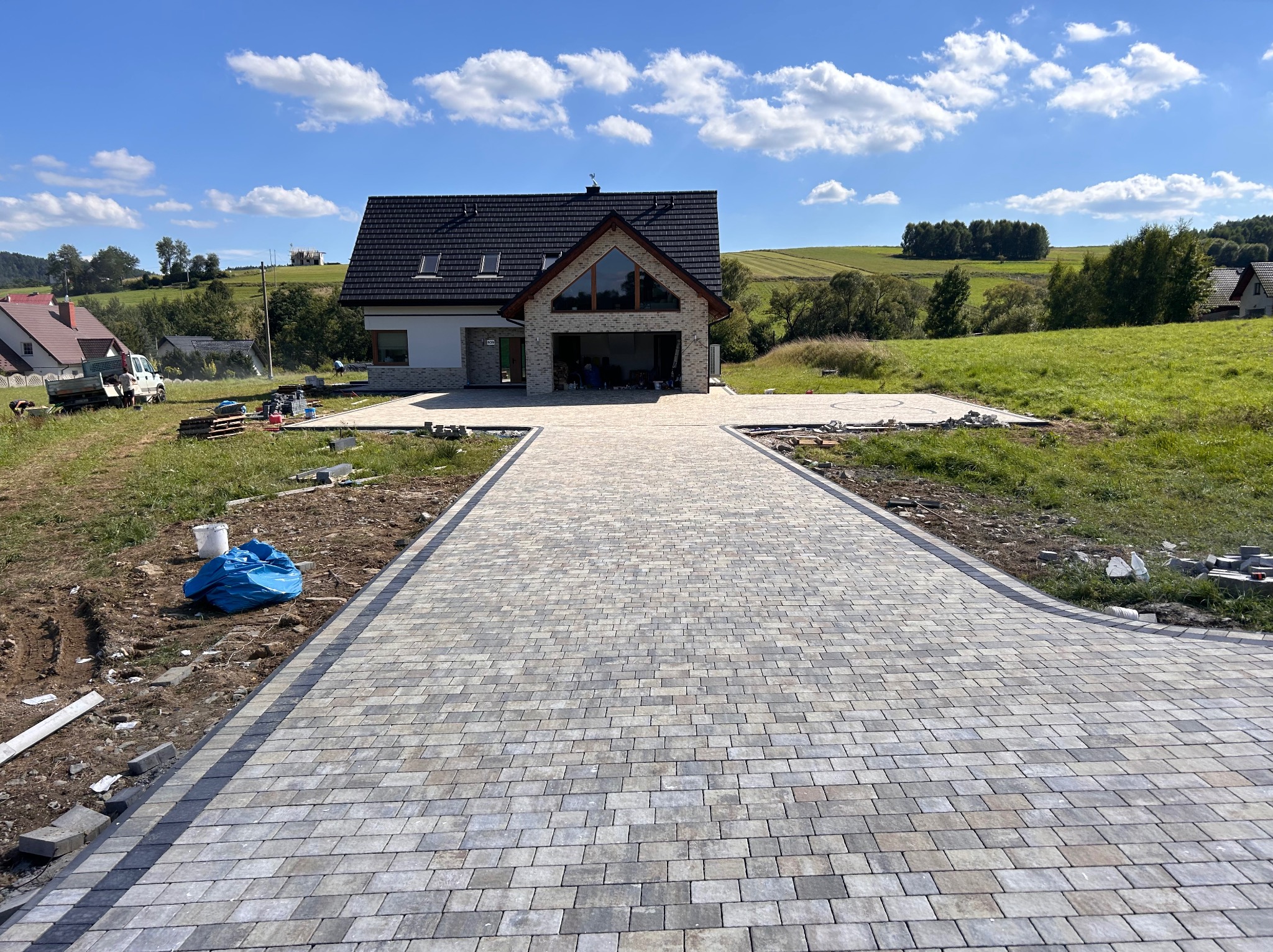 Szeroki podjazd z ułożonej kostki w odcieniach szarości i beżu prowadzący do nowoczesnego domu z garażem, w tle zielone wzgórza i błękitne niebo z chmurami.