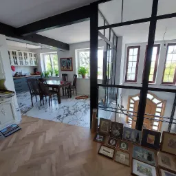 Dwupoziomowe wnętrze domu z widokiem na kuchnię z ciemnymi meblami, drewniany stół z krzesłami, marmurową podłogę oraz przeszkloną ścianę z czarnymi ramami i drewnianymi oknami, a na podłodze leżą...