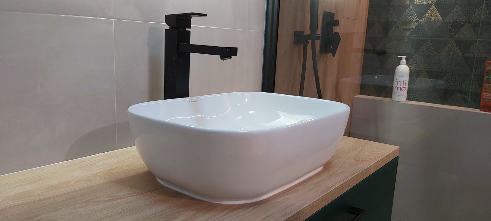 Biała, kwadratowa umywalka nablatowa z czarną armaturą w nowoczesnej łazience z beżowymi płytkami i dekoracyjną ścianą za prysznicem.