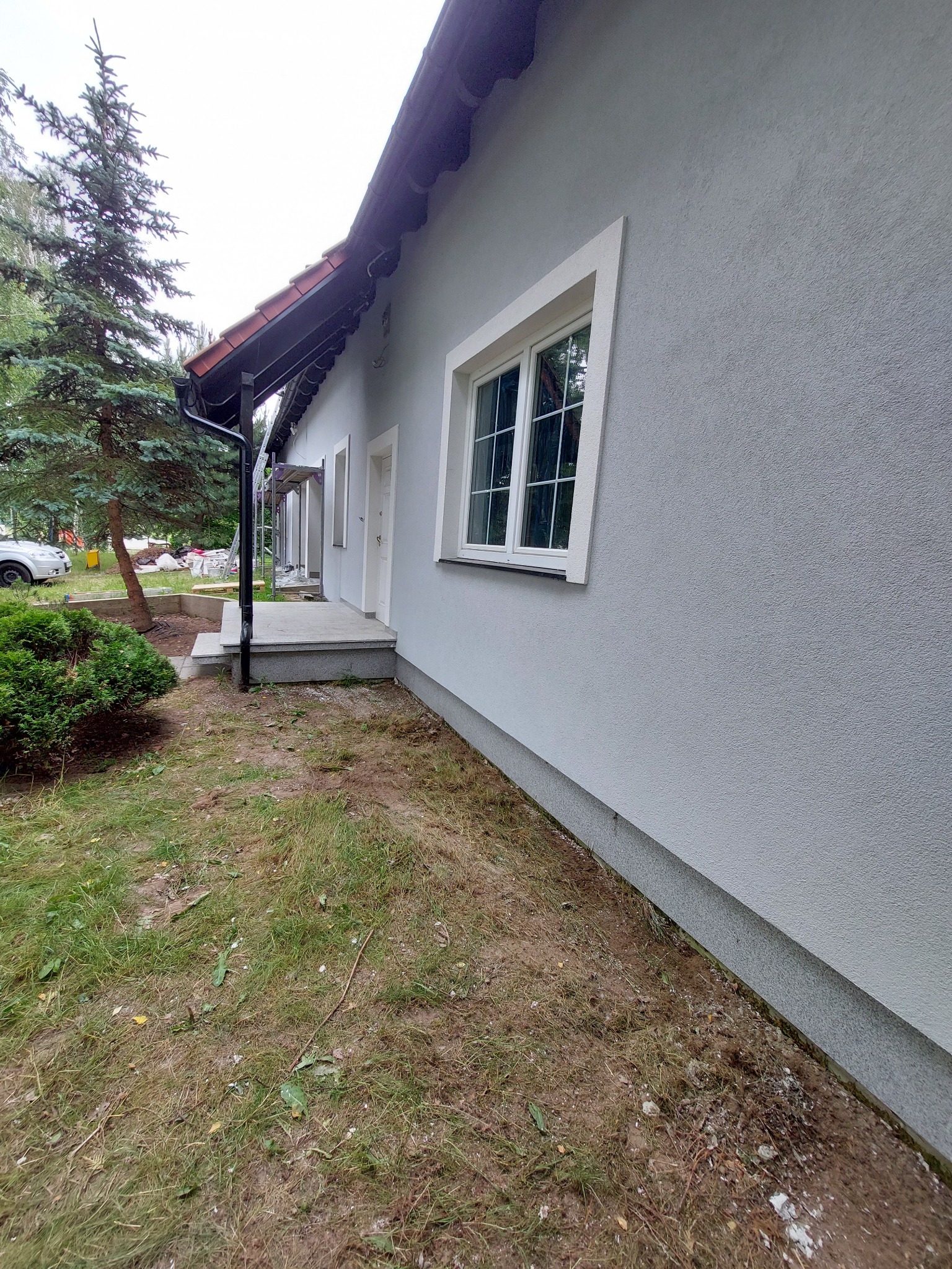 Świeżo otynkowana ściana domu jednorodzinnego w odcieniach szarości z białymi obramowaniami okien, widoczny fragment dachu z czerwoną dachówką i rynną, trawnik z elementami budowy w tle.