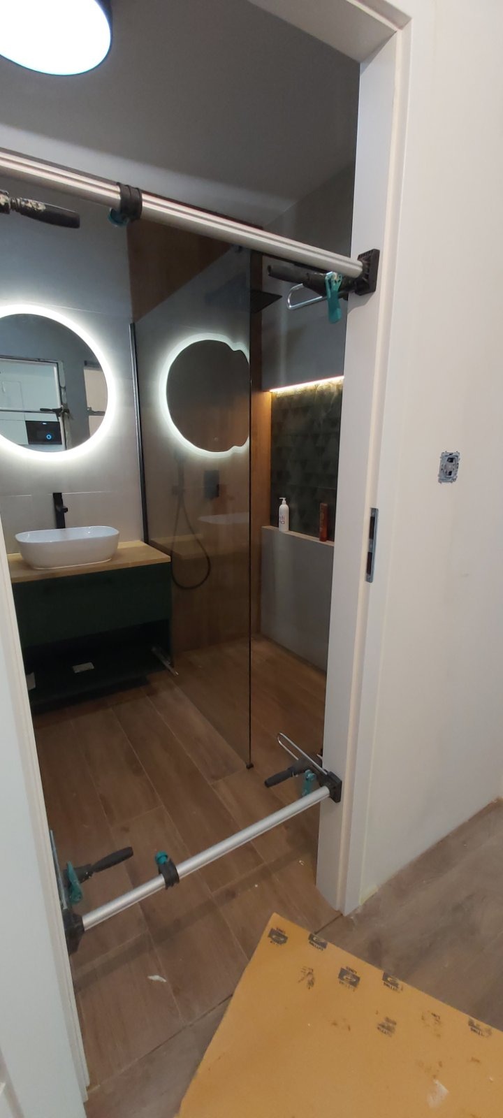 Montaż szklanej kabiny prysznicowej w nowoczesnej łazience z okrągłym lustrem podświetlanym LED i drewnopodobnymi płytkami podłogowymi, widoczne narzędzia montażowe.