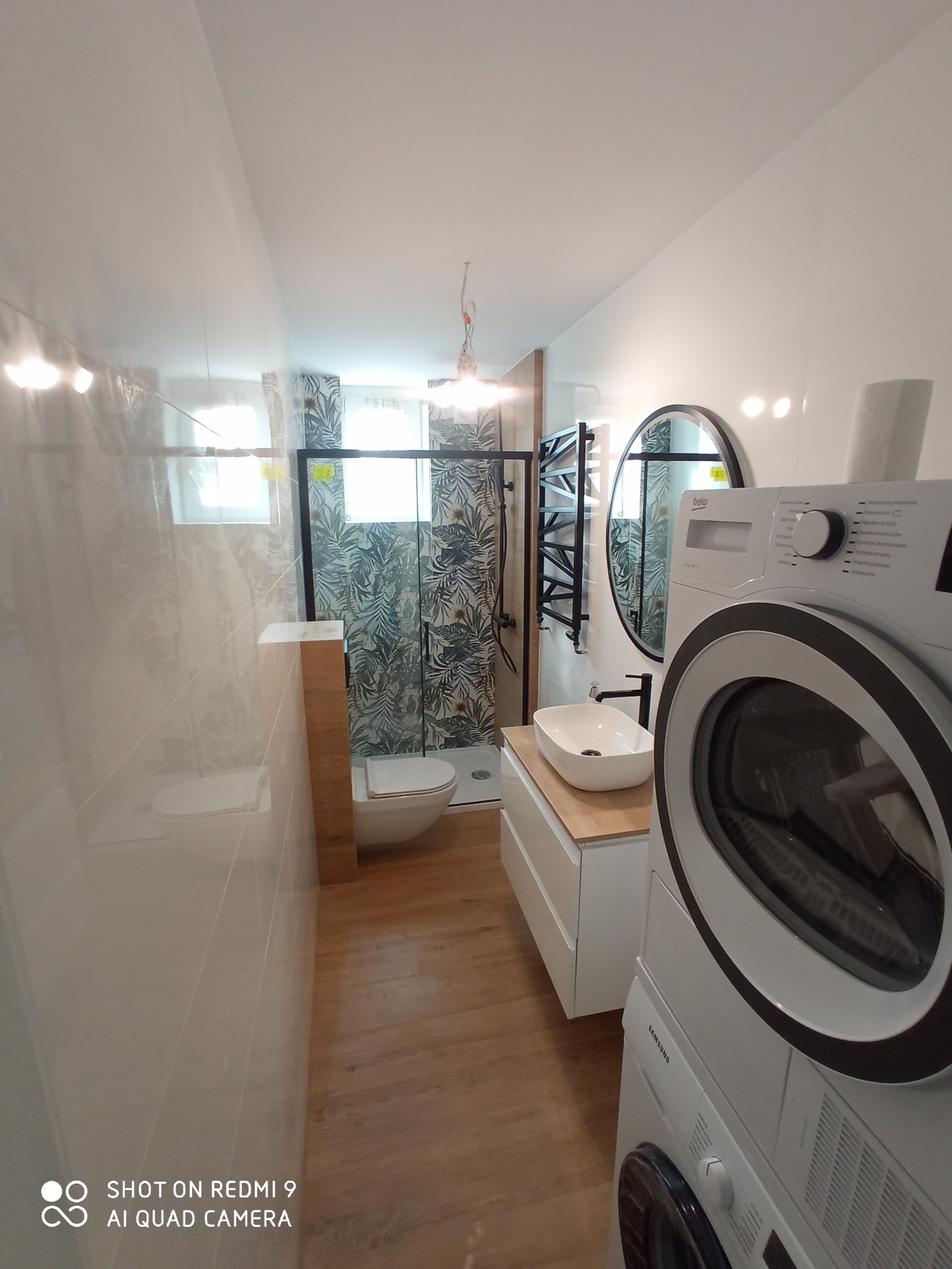 Nowoczesna łazienka z toaletą, kabiną prysznicową z motywem liści, umywalką nablatową na drewnianym blacie, okrągłym lustrem i pralko-suszarką ustawioną jedna na drugiej.