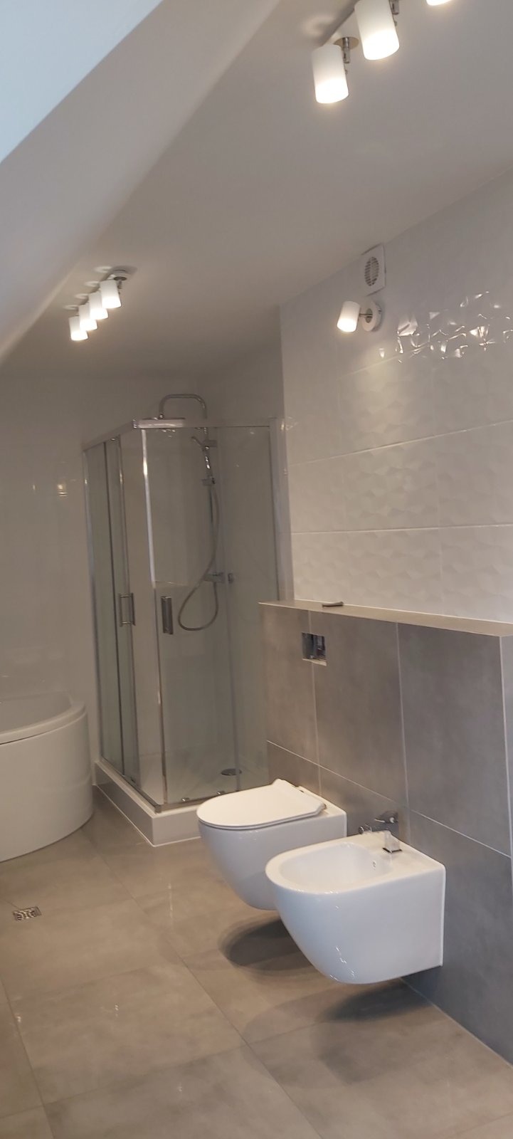 Nowoczesna łazienka na poddaszu z kabiną prysznicową, wiszącą toaletą i bidetem, wykończona szarymi płytkami z subtelnym wzorem i punktowym oświetleniem.