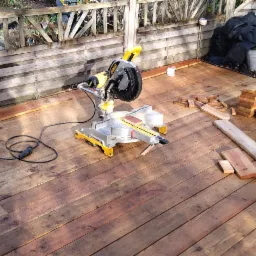 Budowa tarasu z desek: ukończona część podłogi, widoczna piła ukośnica DeWalt, stosy przyciętych desek i drewniana balustrada w tle.