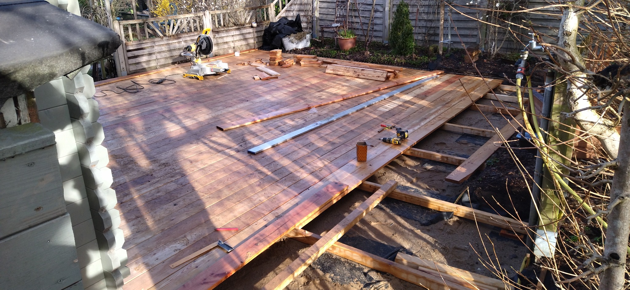 Konstrukcja drewnianego tarasu w trakcie montażu, widoczne legary, częściowo ułożona deska tarasowa, narzędzia: wiertarka, poziomica, piła ukośnica, oraz elementy mocujące.