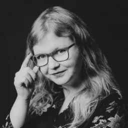 Portret kobiety w okularach, z długimi blond włosami, w ciemnej bluzce w kwiaty, z uniesioną ręką dotykającą oprawki okularów, na czarnym tle.