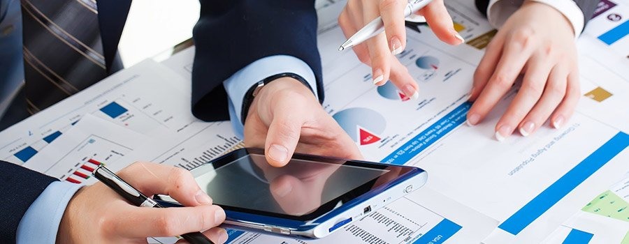 Dwie osoby analizują dane finansowe na tabletach i wykresach; widoczne dłonie z długopisami wskazujące na dokumenty i ekran urządzenia.