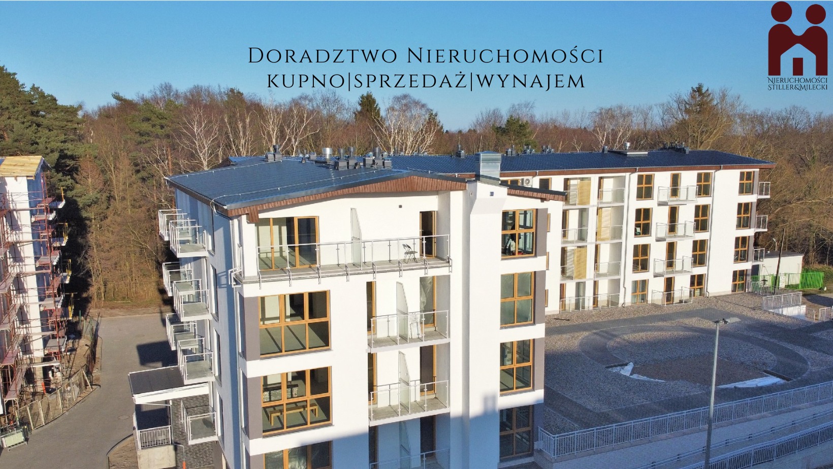 Nowoczesny budynek wielorodzinny z balkonami, częściowo w trakcie wykończenia, widoczny fragment rusztowania z lewej strony, na górze niebieskie niebo i drzewa.