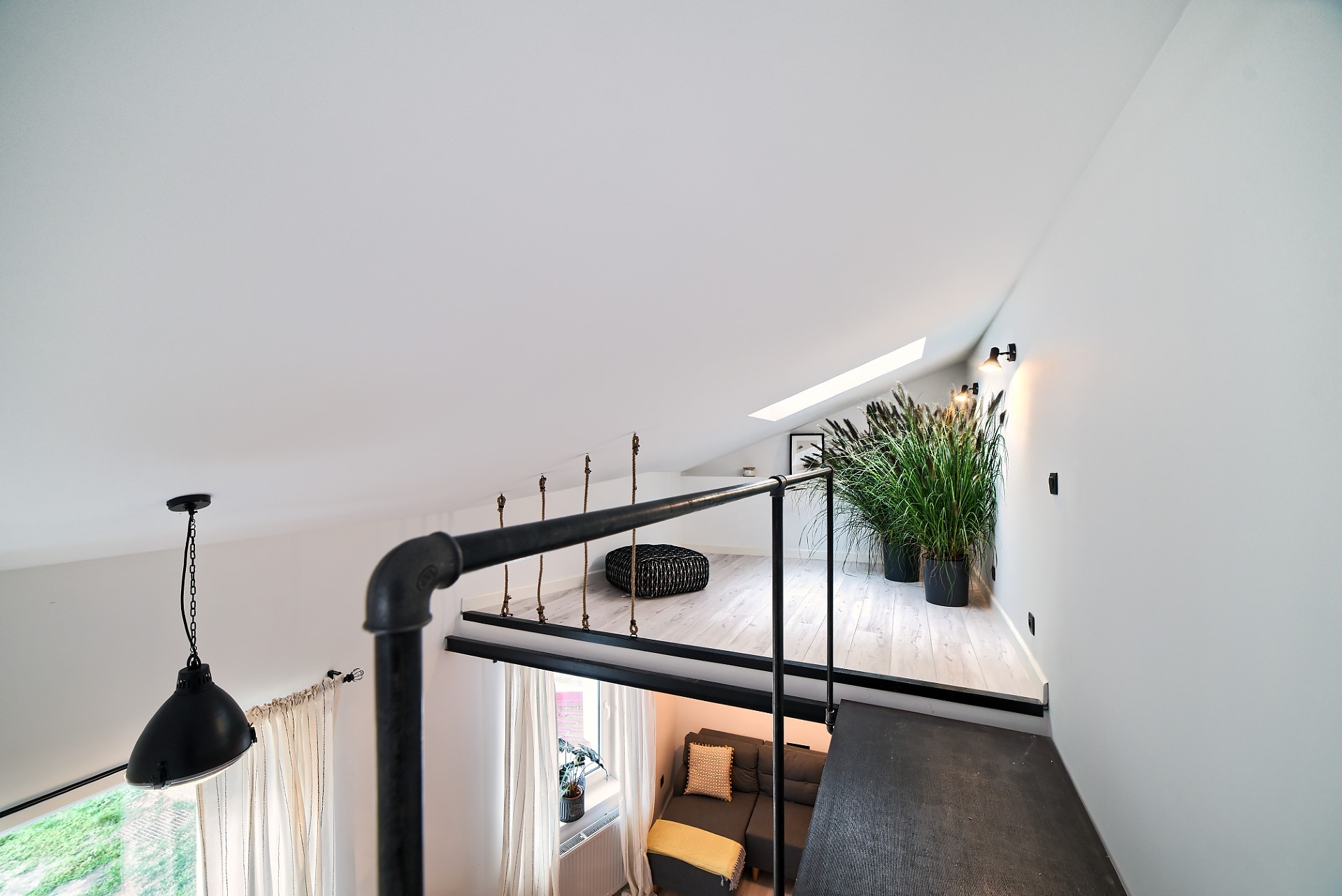 Nowoczesne wnętrze z antresolą, widok z góry na salon z szarą sofą i żółtymi akcentami, antresola z jasną podłogą, pufą i wysokimi trawami w donicach, czarne metalowe barierki i lampa w stylu...