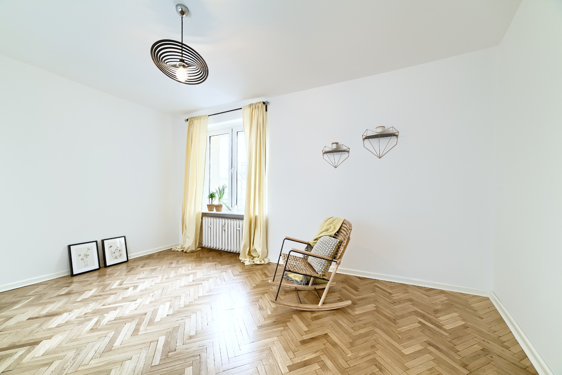 Jasne, puste pomieszczenie w Poznaniu po odświeżeniu, z parkietem w jodełkę, wiklinowym fotelem bujanym z poduszką, nowoczesną lampą i dekoracjami ściennymi.