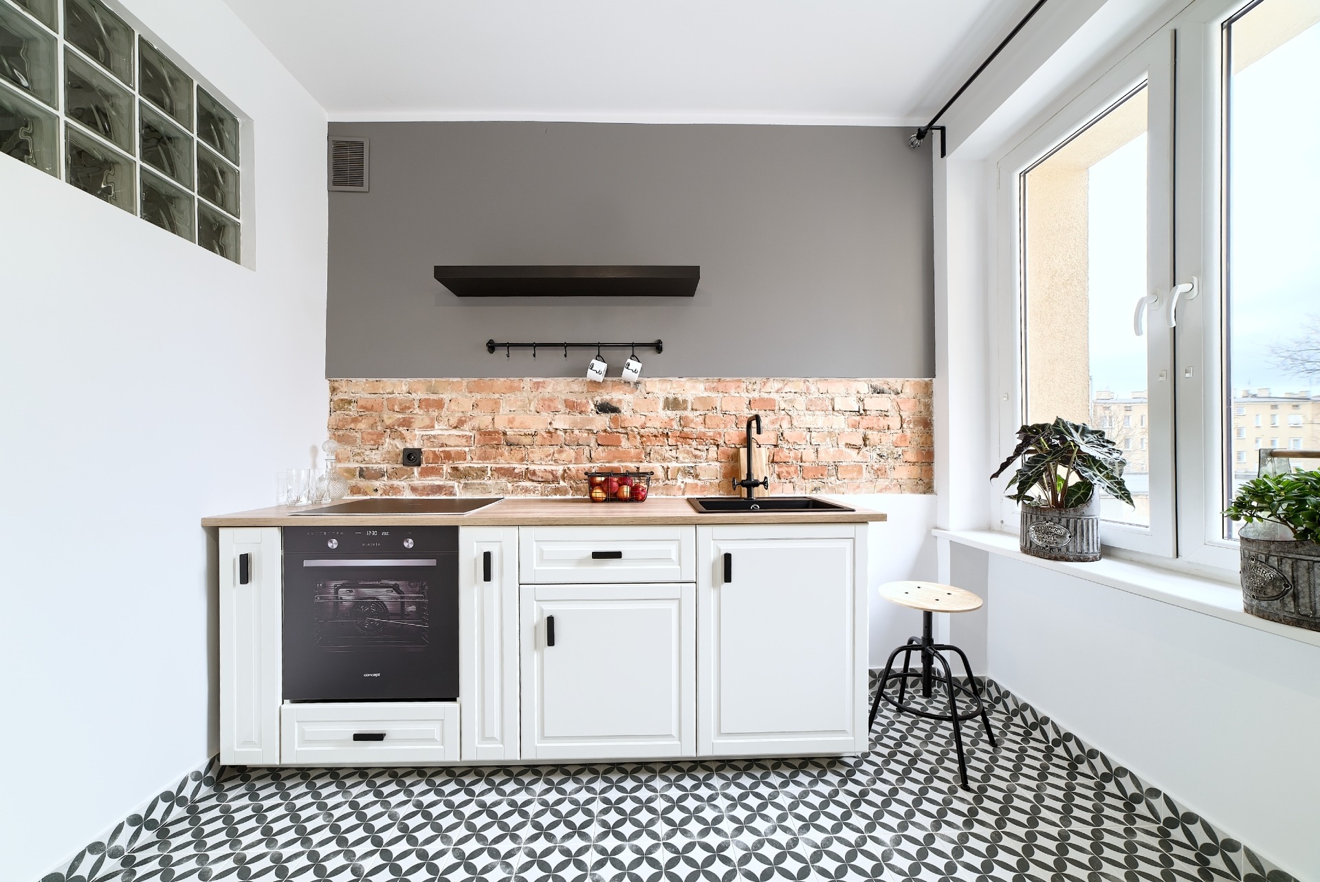 Nowoczesna kuchnia z białymi szafkami, drewnianym blatem, ceglaną ścianą i czarną armaturą, uzupełniona dekoracyjnym stołkiem barowym i roślinami doniczkowymi na parapecie.