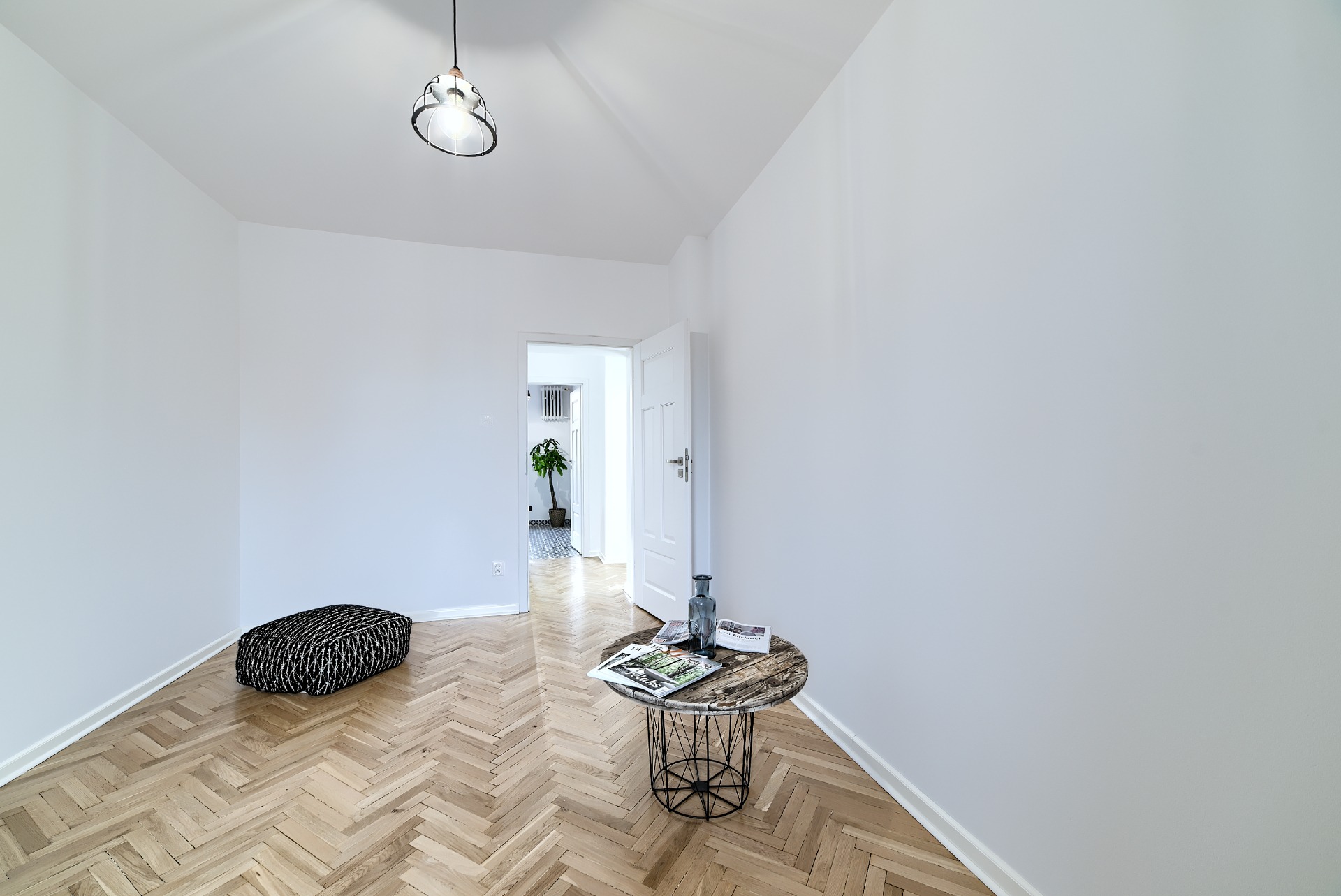 Jasne, puste wnętrze z parkietem ułożonym w jodełkę, z pufą w rogu i okrągłym stolikiem z gazetami oraz wazonem, widok na otwarte drzwi do kolejnego pomieszczenia z rośliną doniczkową.