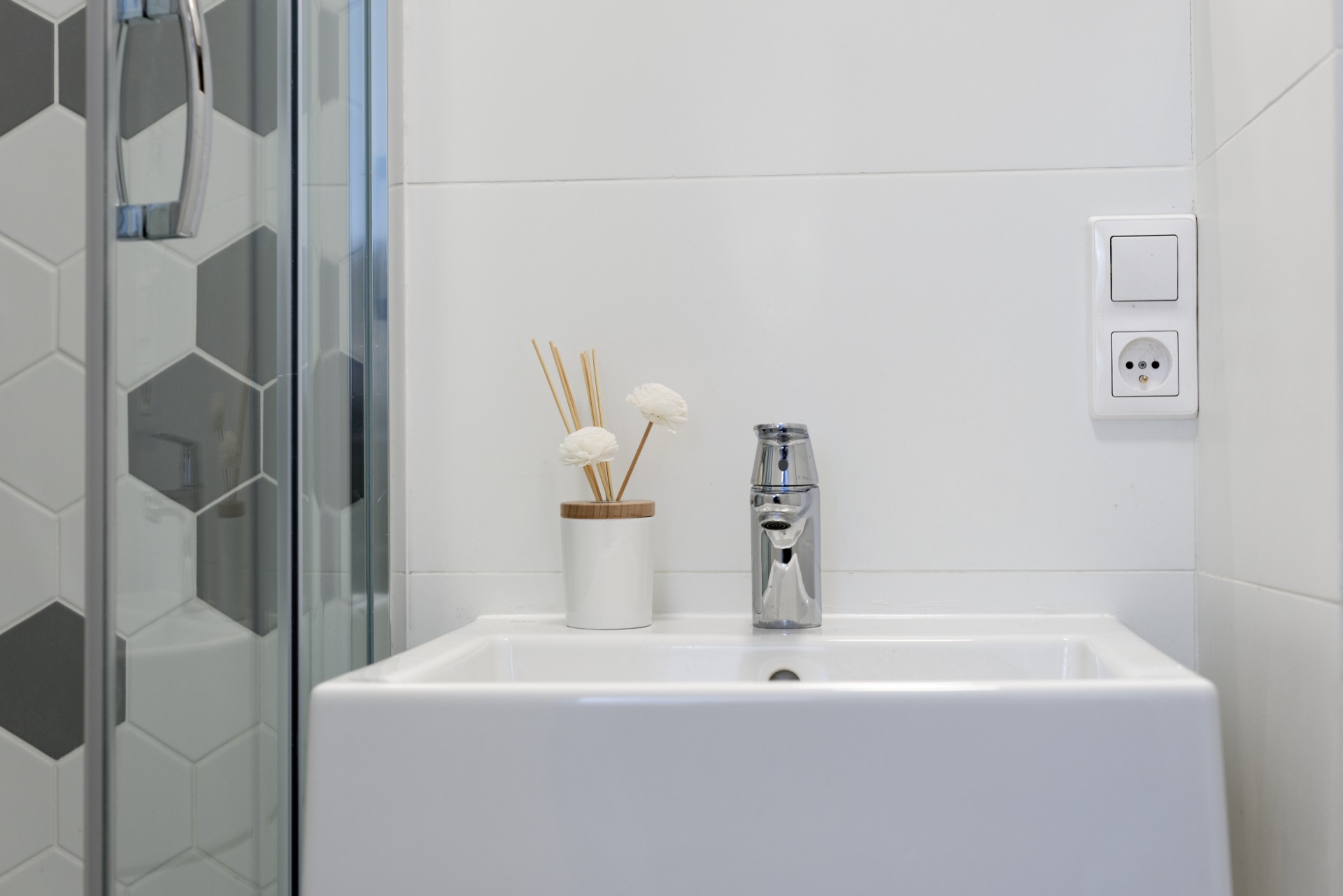 Biała umywalka nablatowa z chromowaną baterią, dyfuzor zapachowy z białymi kwiatami i szklana kabina prysznicowa z heksagonalnymi płytkami w odcieniach szarości.