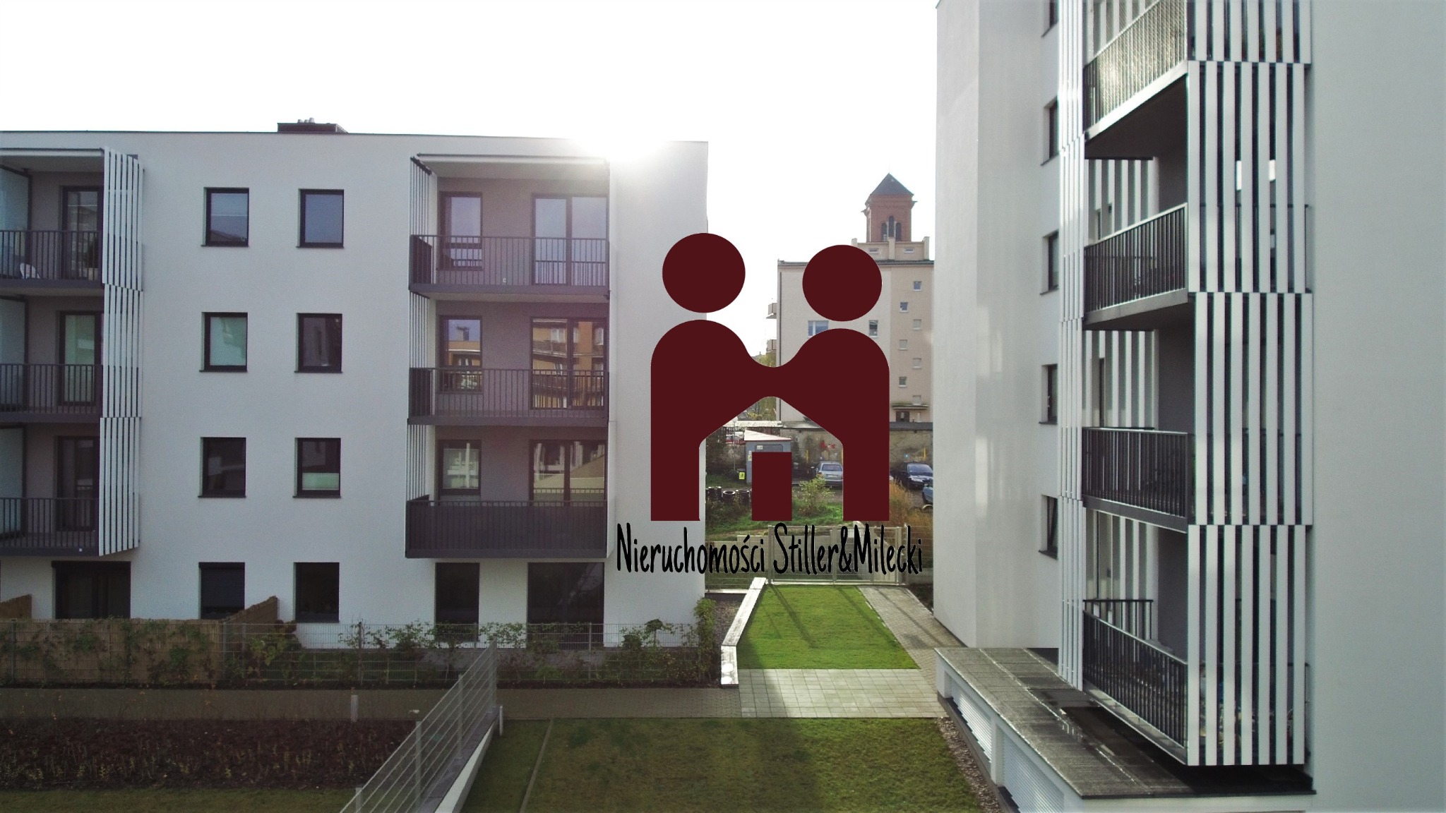 Nowoczesny budynek apartamentowy z balkonami i minimalistyczną elewacją, widoczny z perspektywy trawnika, z nałożonym logo firmy.