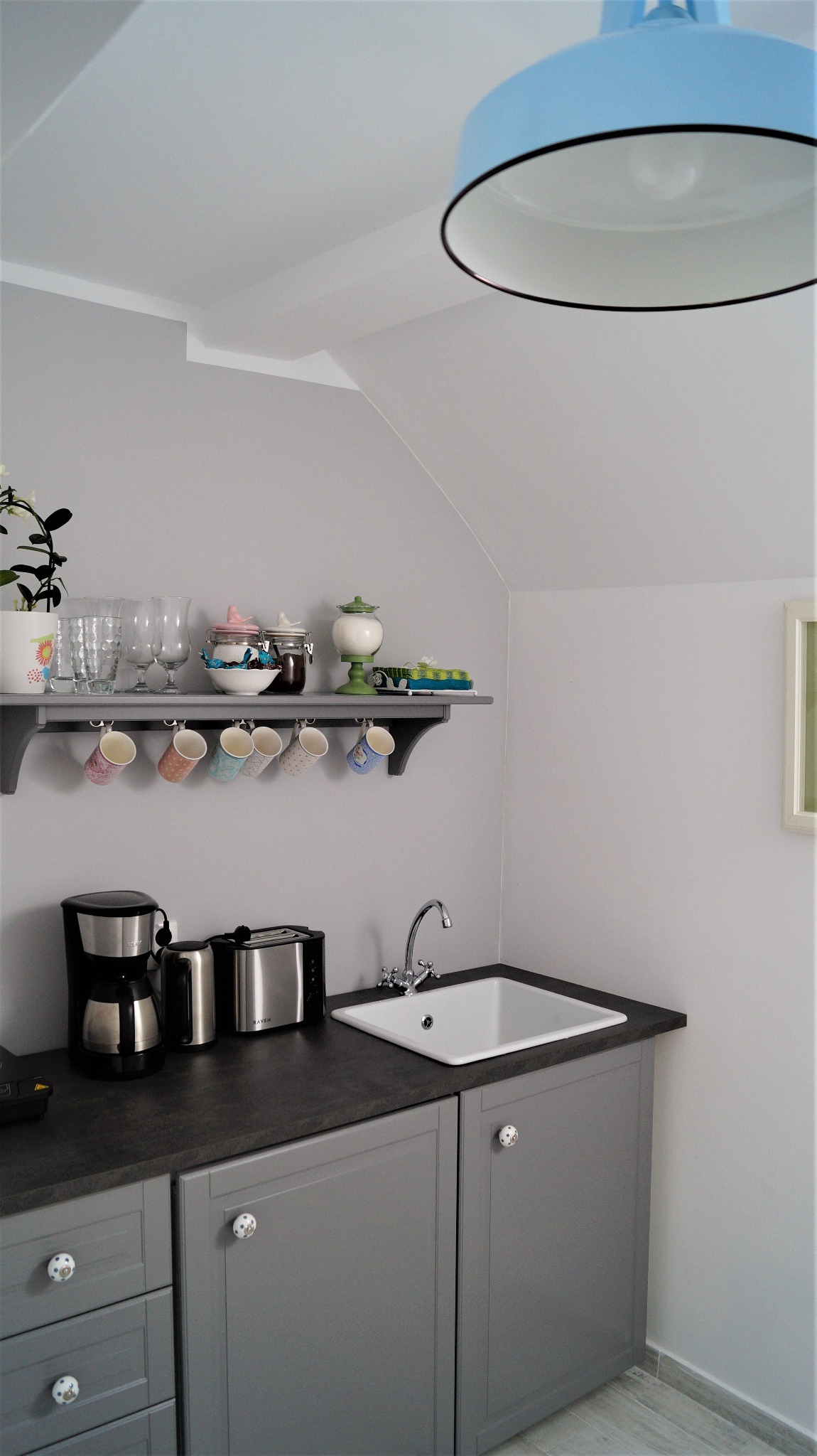 Szara kuchnia w skandynawskim stylu z białą ceramiczną umywalką, chromowaną baterią, czarnym blatem, szarymi szafkami z ozdobnymi gałkami, półką z naczyniami i błękitną lampą.