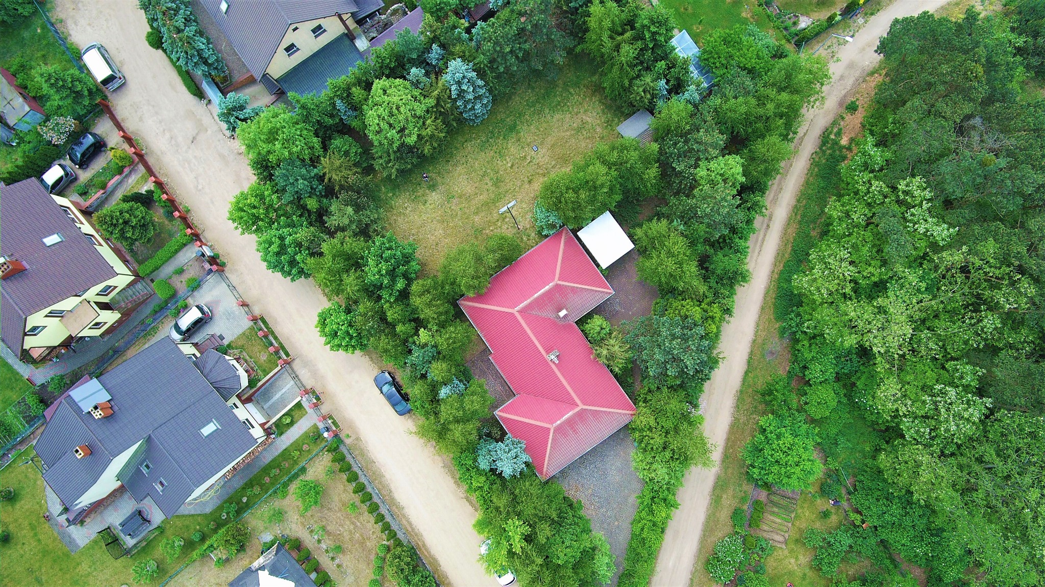 Zdjęcie z lotu ptaka przedstawia dom z czerwonym dachem otoczony zielenią, w pobliżu piaszczysta droga i inne zabudowania.