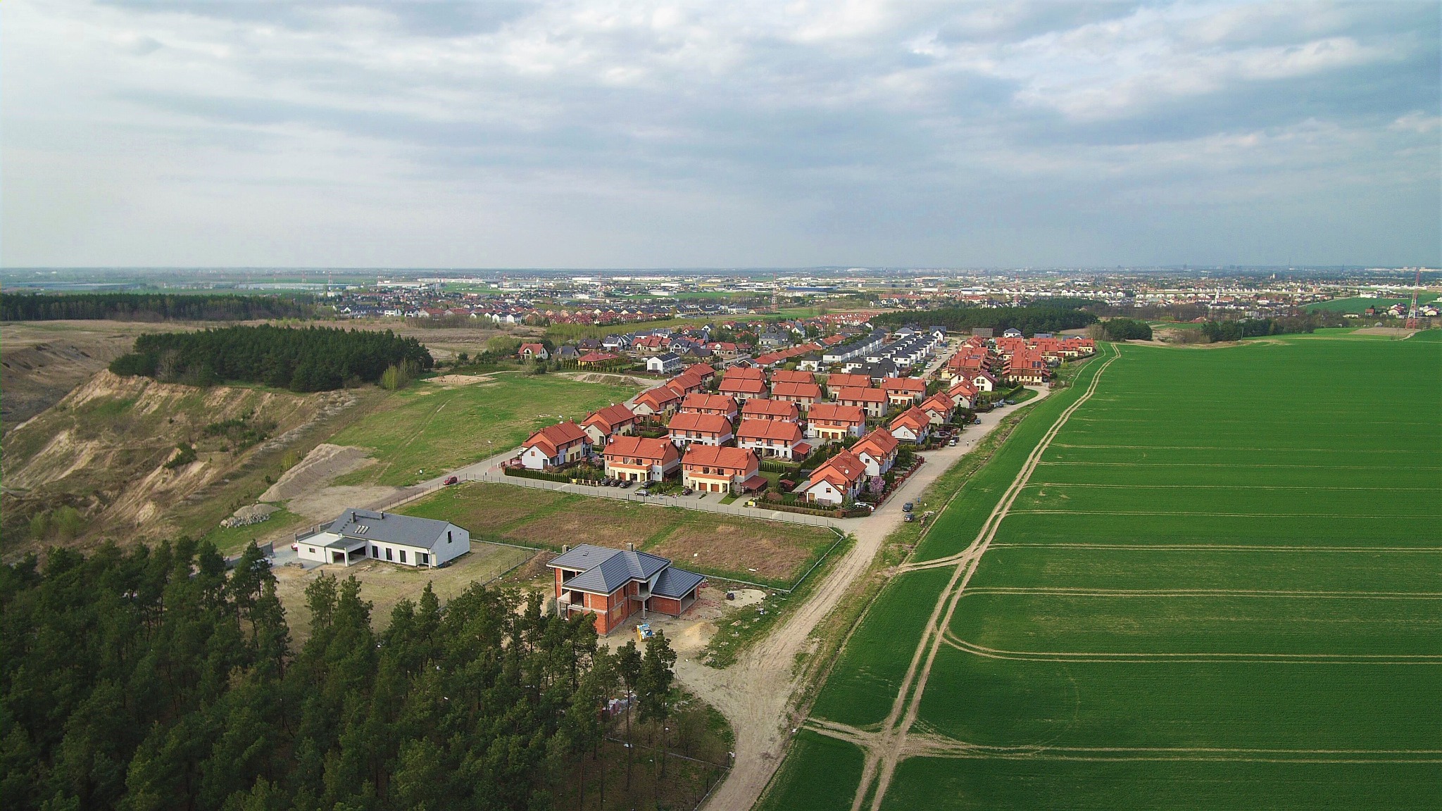 Widok z lotu ptaka na osiedle domów jednorodzinnych z czerwonymi dachami, z jednym domem w trakcie budowy, obok zielone pole uprawne i las, w tle zabudowa miejska.