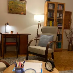 Gabinet z drewnianym biurkiem, lampką, krzesłem, fotelem z poduszką, regałem z książkami, okrągłym stolikiem z przyborami do pisania, zegarem i podkładką z kartkami.