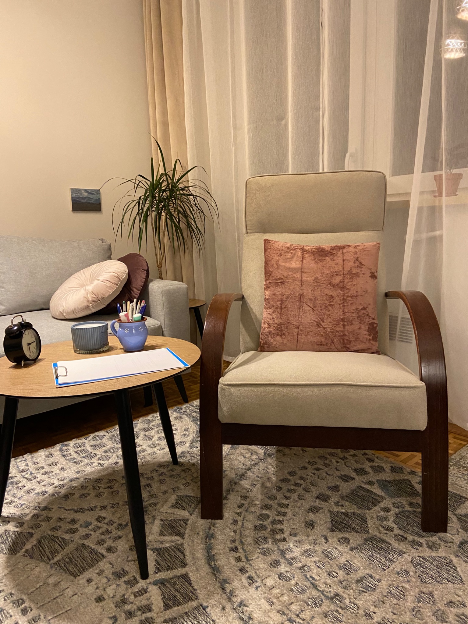 Gabinet psychoterapeutyczny: fotel z poduszką, stolik z notatnikiem i przyborami, sofa z okrągłymi poduszkami, roślina doniczkowa, dywan w geometryczne wzory.