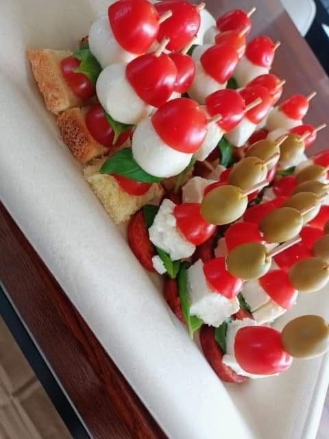 Dekoracyjne przekąski: koreczki z pomidorkami cherry, mini mozzarellą, oliwkami i listkami bazylii, ułożone na grzance z ciabatty i serwowane na białej serwetce.