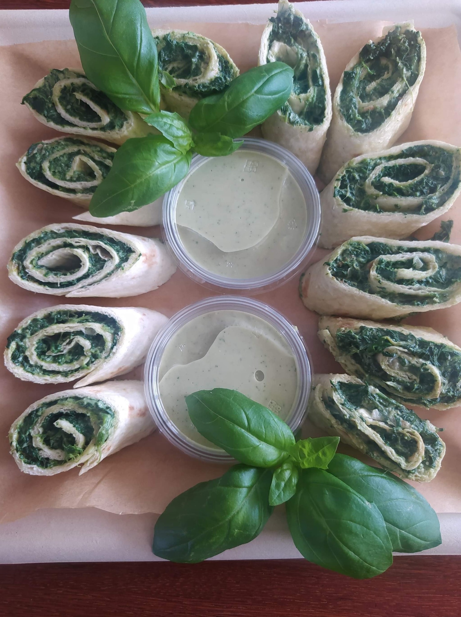 Roladki z tortilli wypełnione szpinakiem, ułożone wokół dwóch pojemników z zielonym sosem, udekorowane świeżą bazylią na papierze.