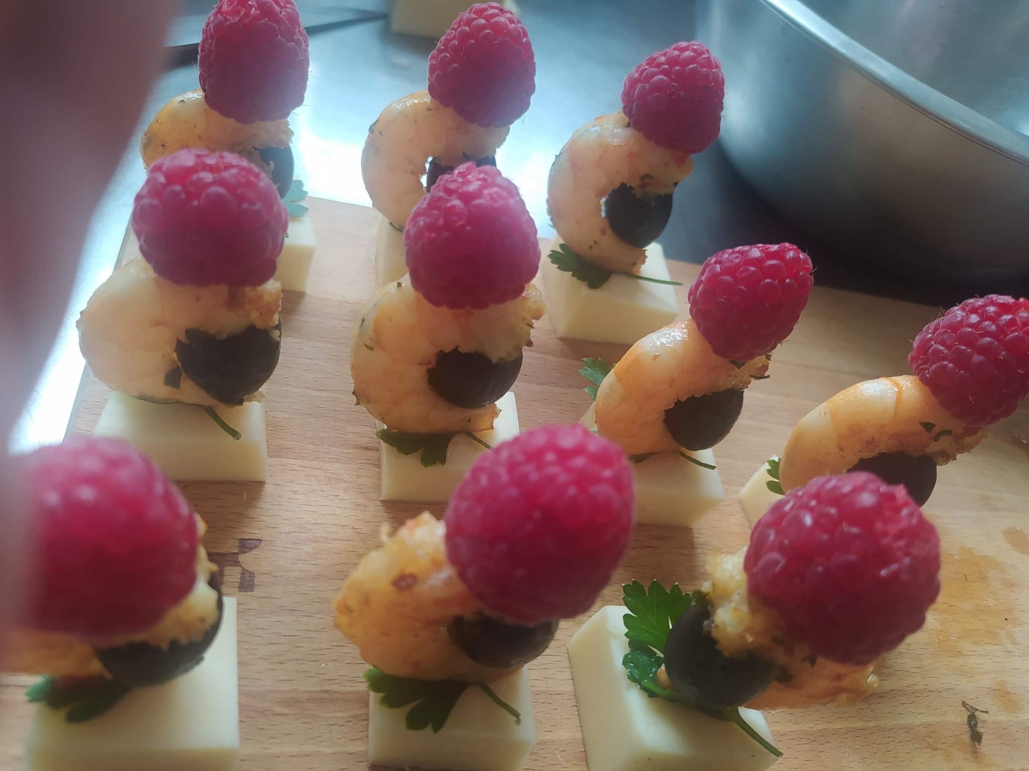 Apetyczne koreczki na drewnianej desce: kostka sera, krewetka, oliwka i malina, udekorowane listkiem pietruszki.