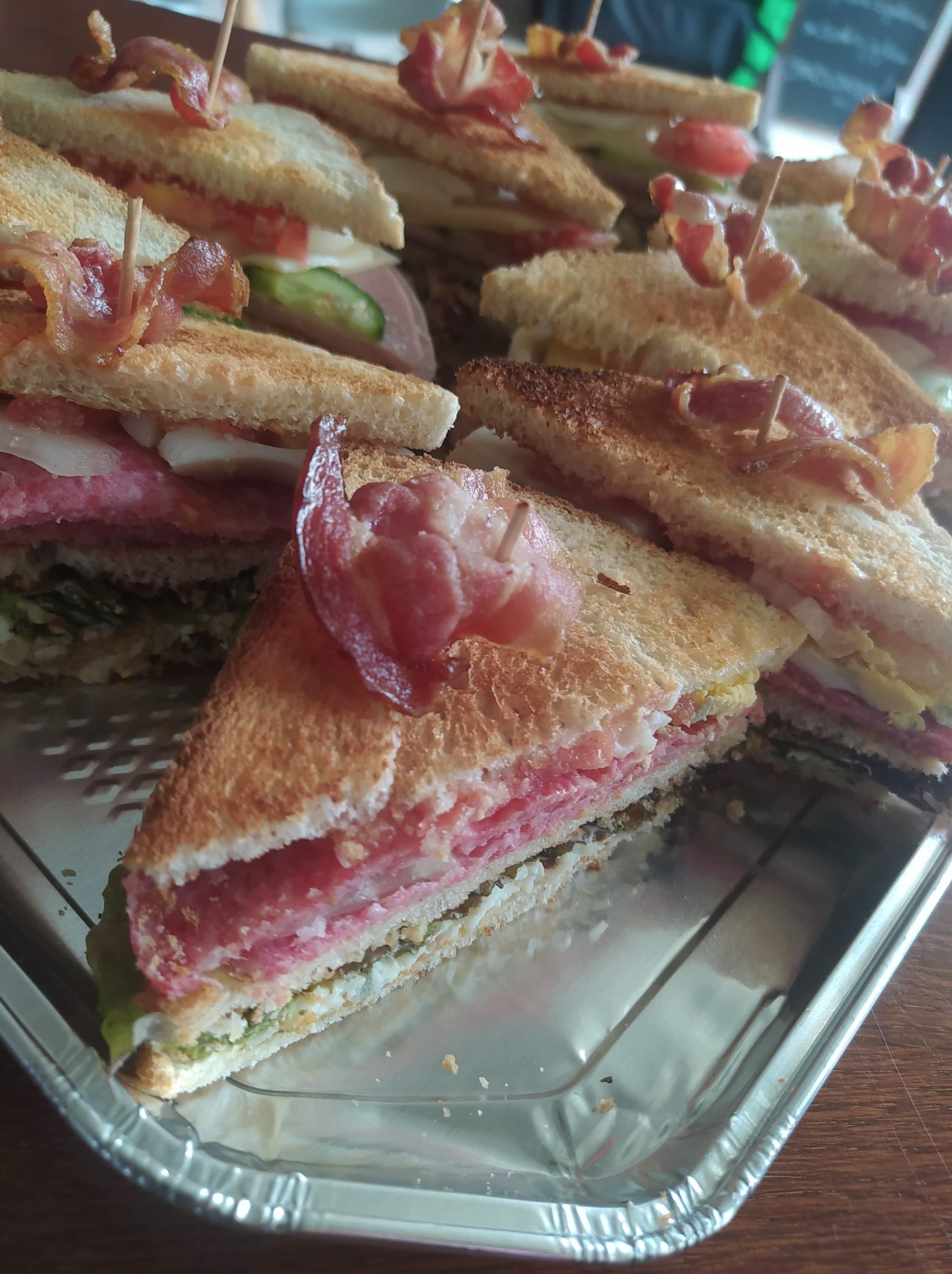 Kilkanaście trójkątnych kanapek na srebrnej tacy, każda z boczkiem przymocowanym wykałaczką, widoczne warstwy sałaty, wędliny, jajka i pomidorów.