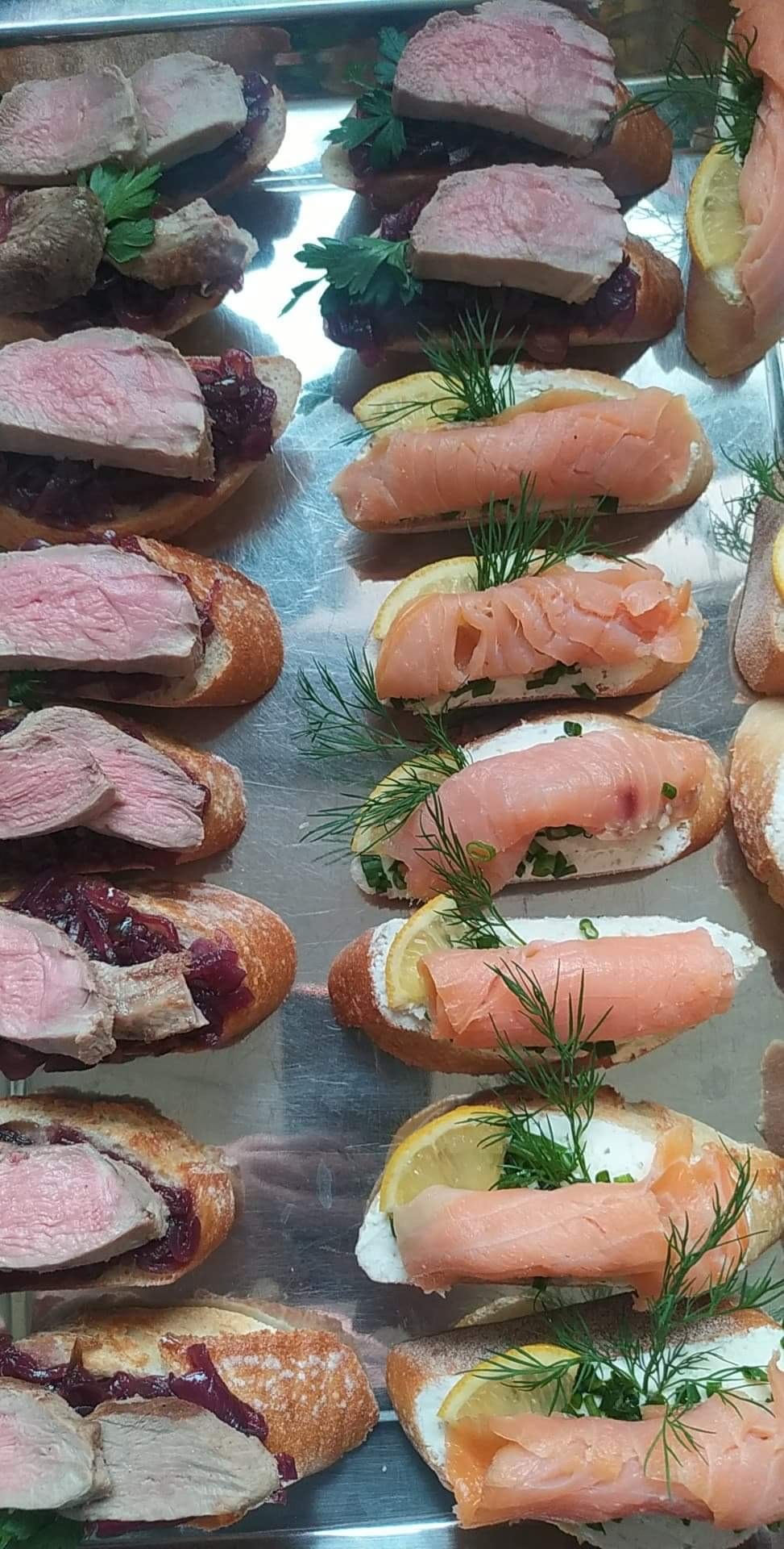 Metalowa taca z różnorodnymi kanapkami: jedne z pieczonym mięsem i czerwoną konfiturą, inne z łososiem, kremowym serem, cytryną i koperkiem.